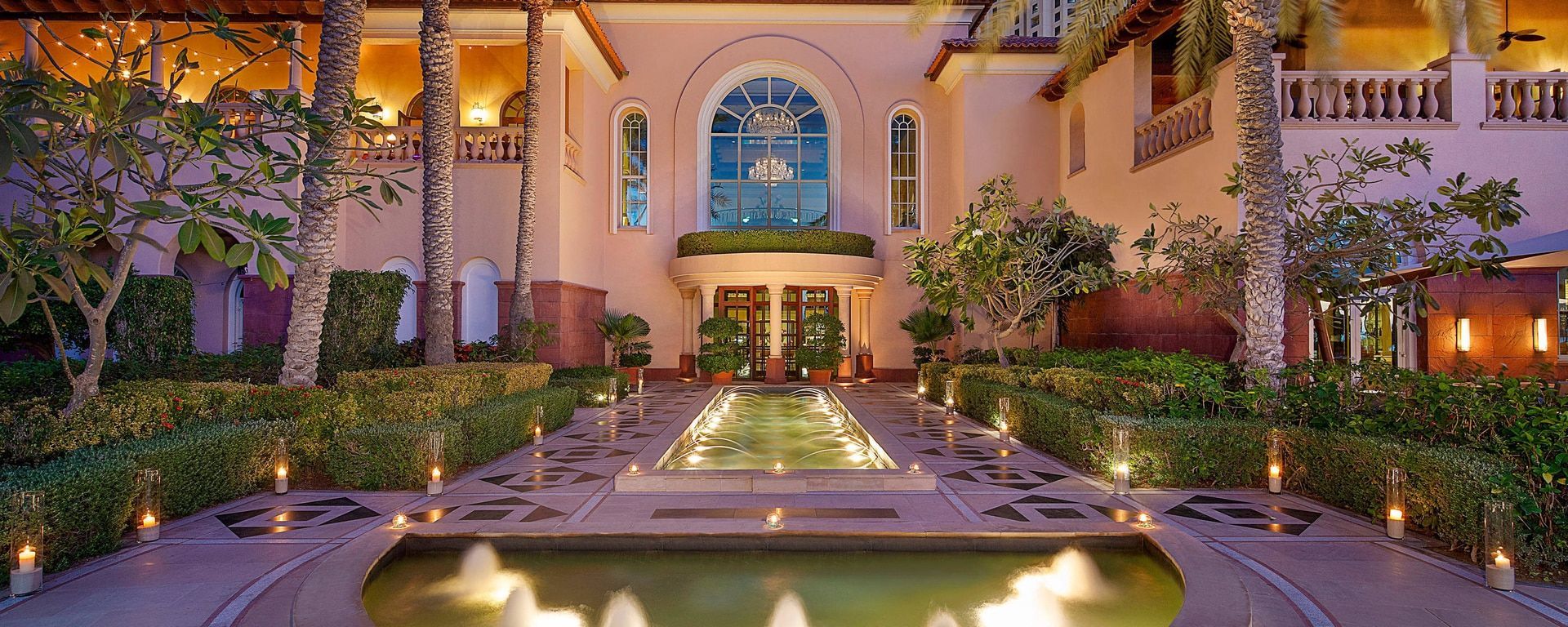 Exterior of a luxurious building with a water feature and lit pathways, surrounded by greenery.