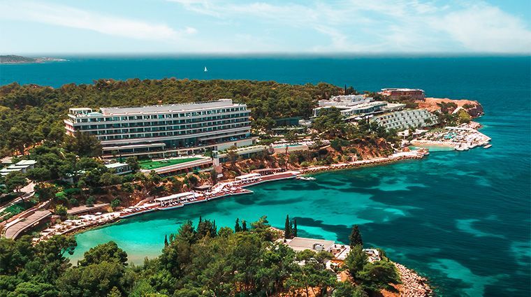 Aerial view of a luxury hotel complex nestled in a forested coastal peninsula with a bright turquoise cove and blue sea.Four Seasons Astir Palace Hotel Athens