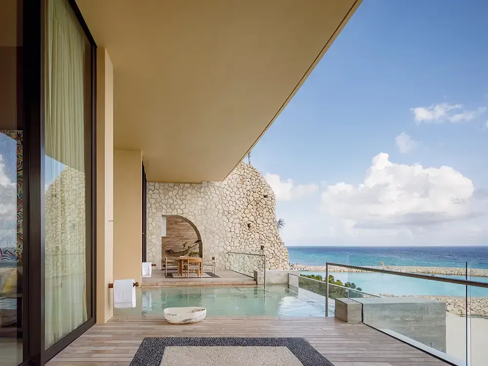 A wooden balcony with a private infinity pool overlooking the ocean, framed by stone walls under a clear blue sky.