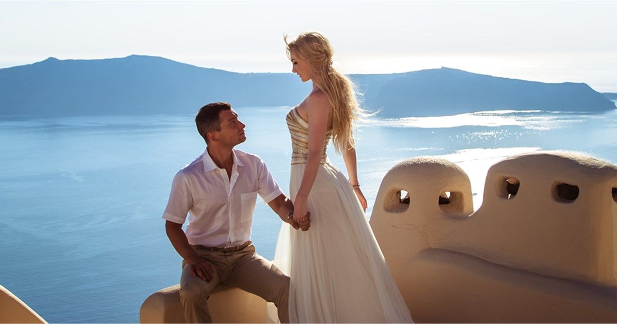 A person in a white dress stands while another person sits nearby, against a backdrop of a blue sea and Greek architecture.