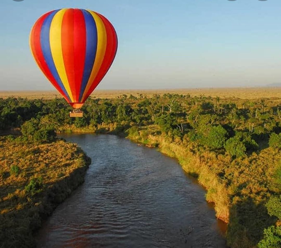 Hot air balloon floats over a river and African savanna. Balloon is red, yellow and blue.