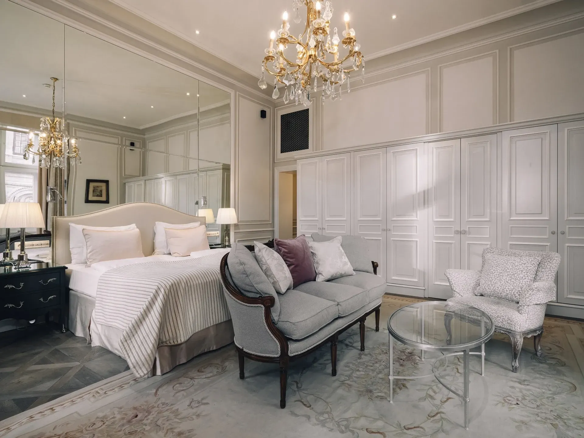 Elegant white bedroom with chandelier, mirrored walls, tufted bed, gray chaise, and glass tables Hotel Sacher Vienna
