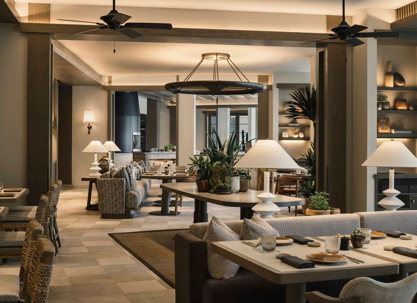 A dimly lit, upscale restaurant interior with neutral-toned seating, wooden tables, warm lamps, and a circular chandelier.