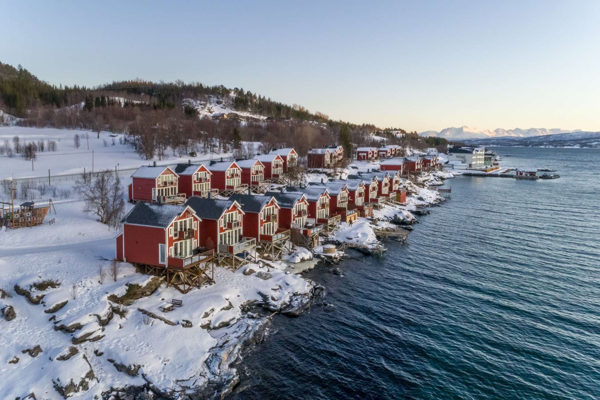A row of red cabins sits on a snowy, rocky shoreline beside a blue fjord under a clear sky. Malangen Resort
