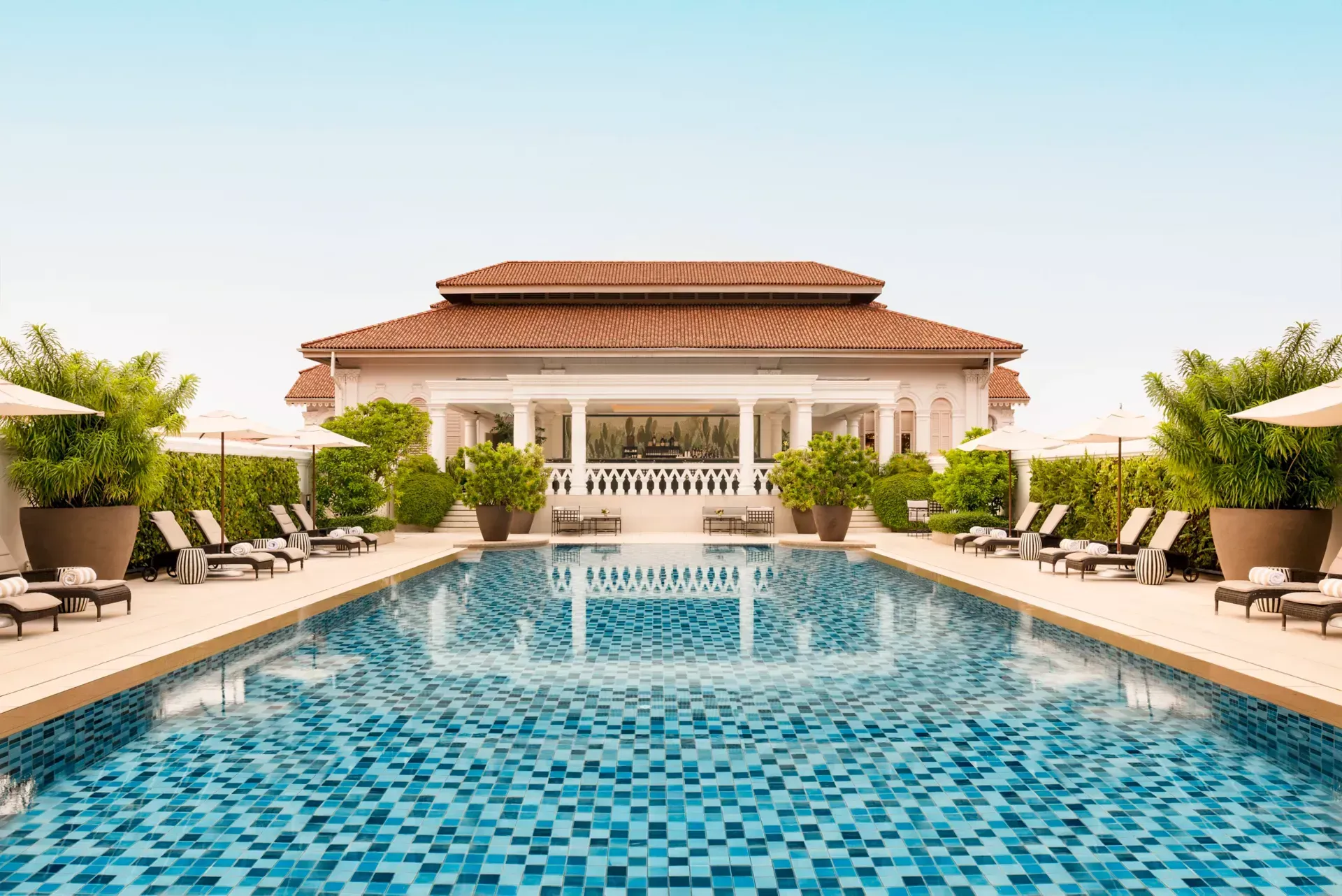 An outdoor swimming pool with blue mosaic tiles, lined with lounge chairs, potted plants, and a building in the background.