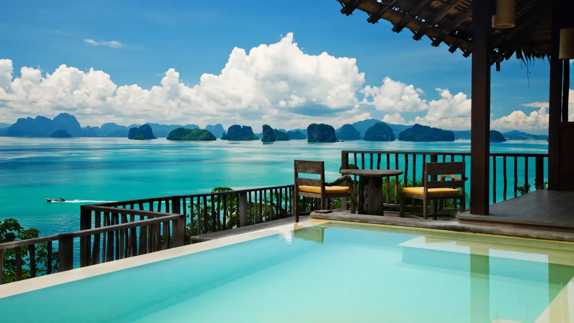 An infinity pool overlooks a calm turquoise ocean with lush, mountainous islands under a bright, cloudy blue sky.