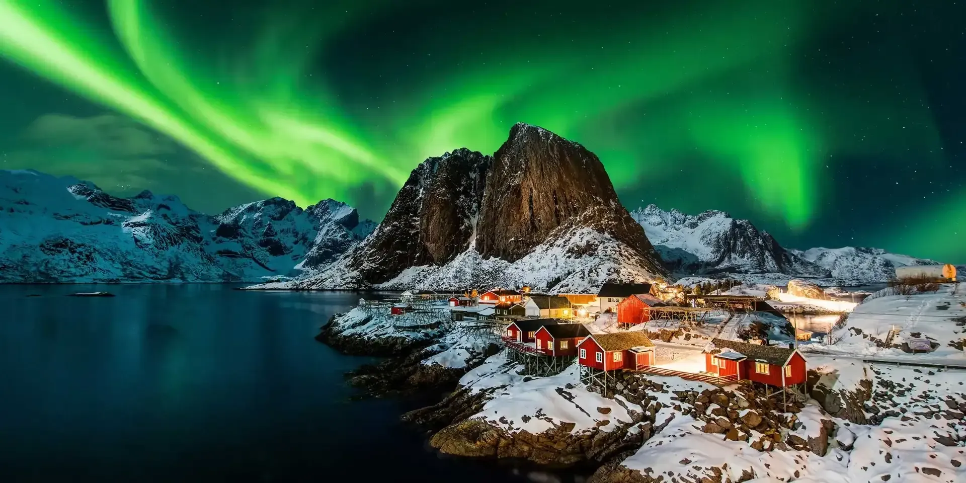Vibrant green aurora borealis illuminates a snowy, mountainous landscape featuring small red cabins along a dark bay.