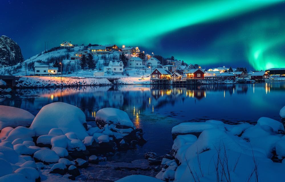 Bright green aurora borealis shines over a snowy village and its reflection in a calm, dark bay at night. Snowhotel Kirkenes, Kirkenes