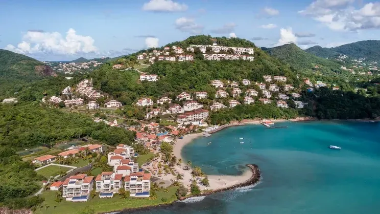 Aerial view of a coastal resort with white buildings nestled on a green hillside next to a sandy beach and turquoise sea. Windjammer Landing 