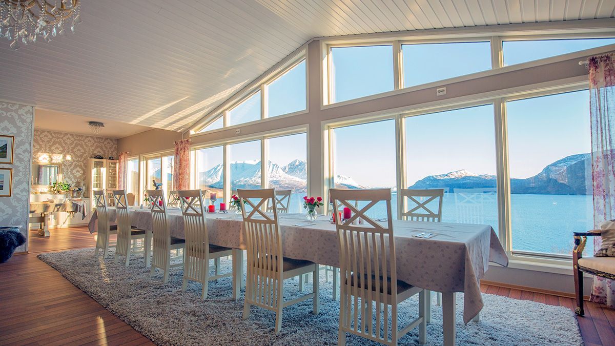 A long dining table set for a meal in a bright, modern room with floor-to-ceiling windows overlooking a snowy mountain bay. Arctic Panorama Lodge, Uløya
