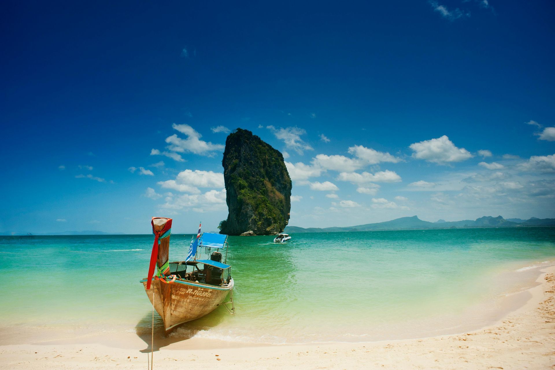 Boat on a white sand beach, turquoise water, large rock island under a bright blue sky. Thailand