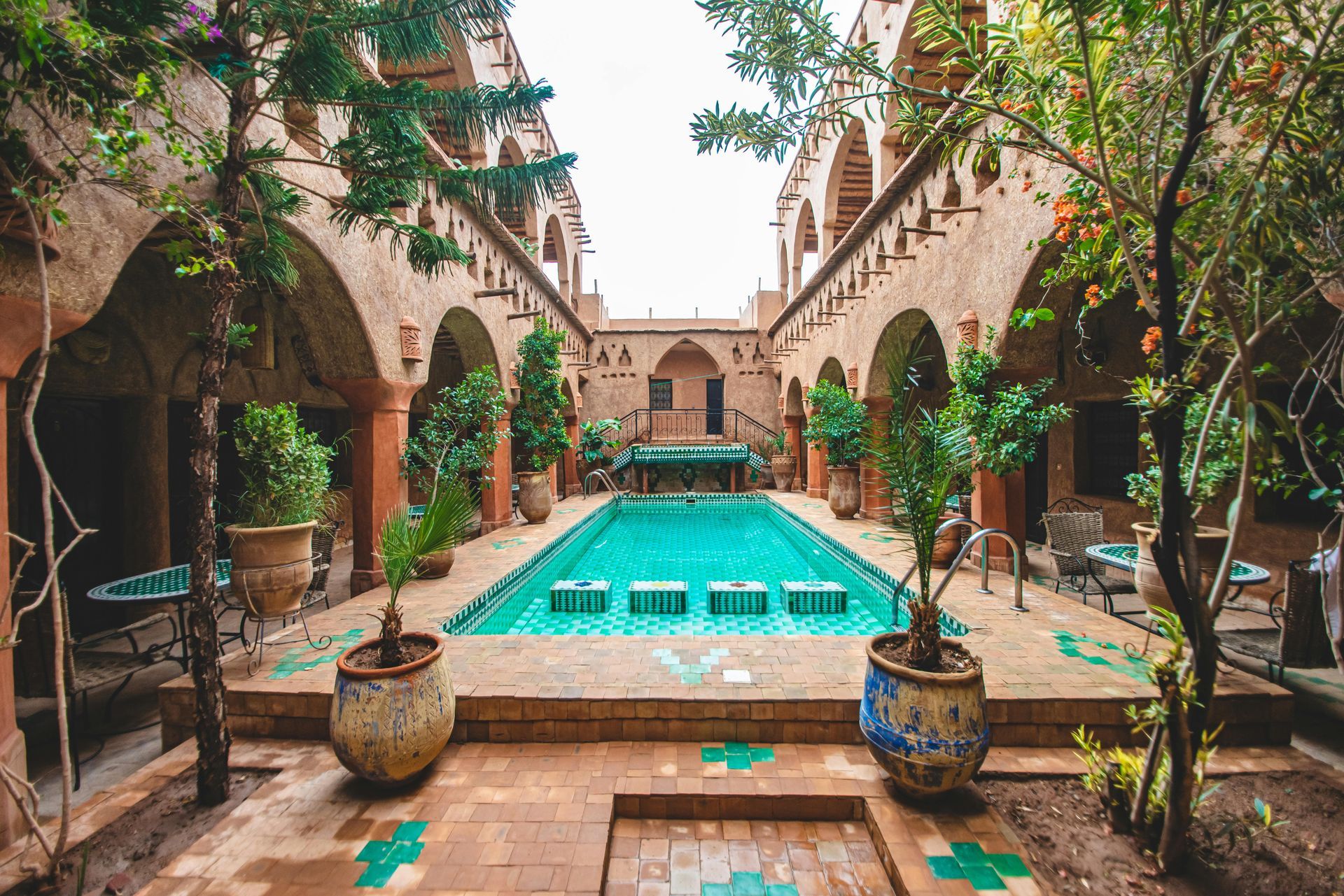 A bright turquoise swimming pool surrounded by terracotta-colored arches, potted plants, and patterned tile accents.