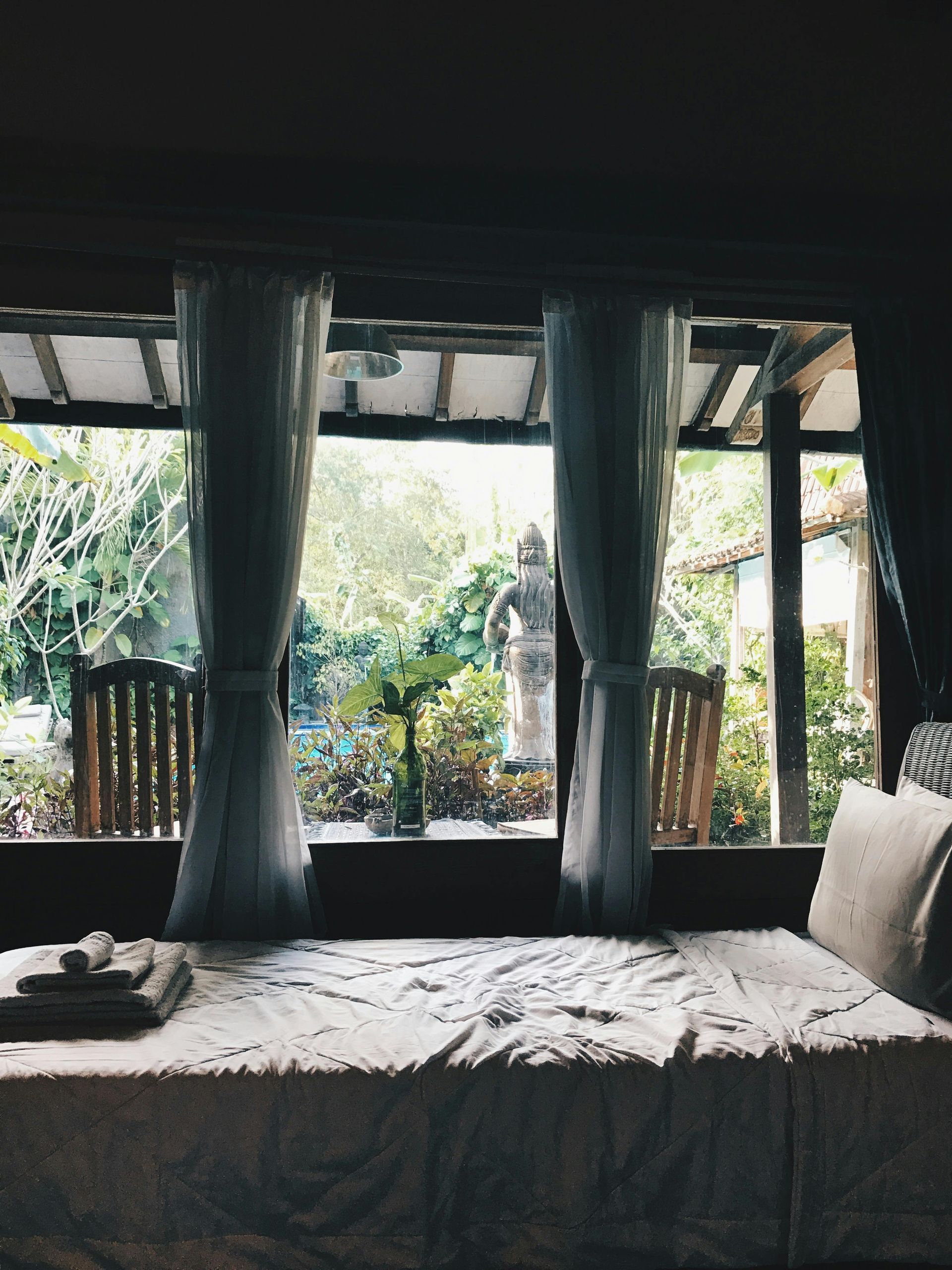 Bedroom with a bed, sheer curtains, and a view of a lush green garden through large windows.
