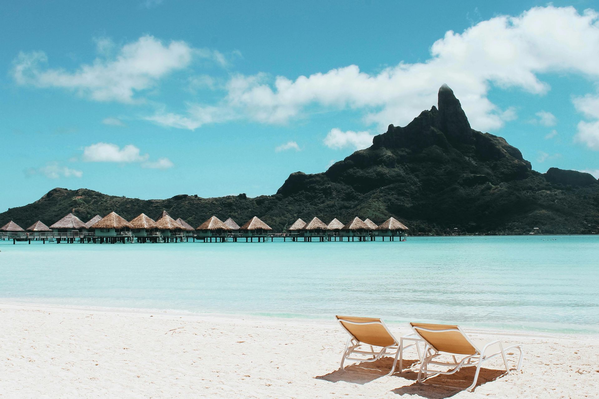 Two lounge chairs face a calm turquoise ocean with overwater bungalows and a mountain under a blue sky. Bora Boa