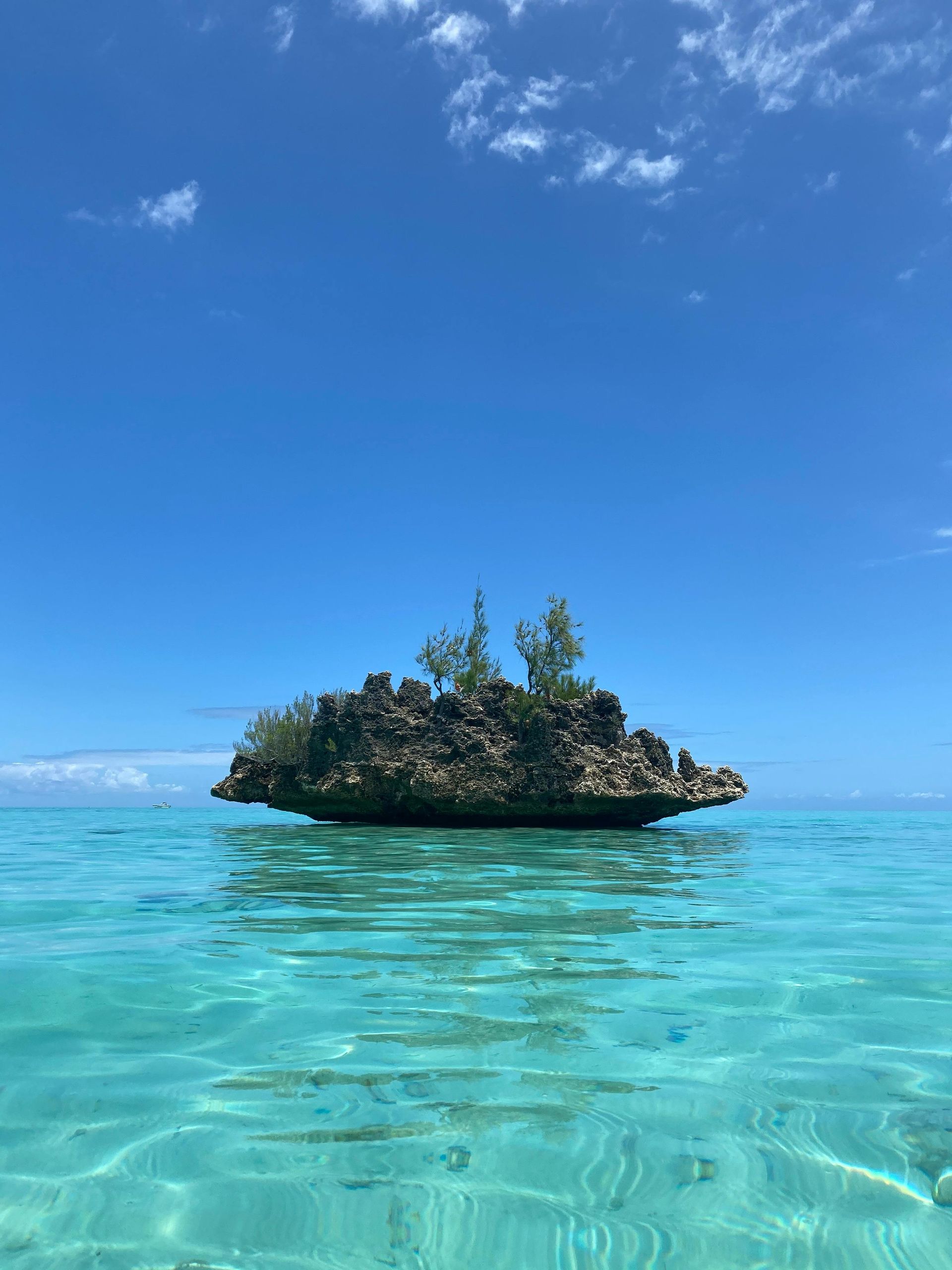 A small, rocky island with sparse vegetation sits in the middle of calm, crystal-clear turquoise water under a blue sky.