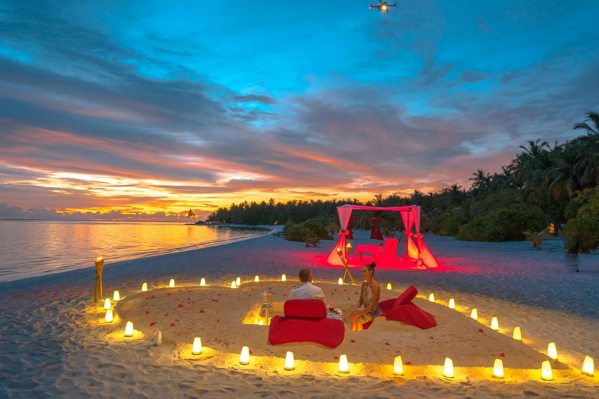 Romantic beach dinner setup with heart-shaped candles, red decor, and sunset backdrop. Soneva Jani
