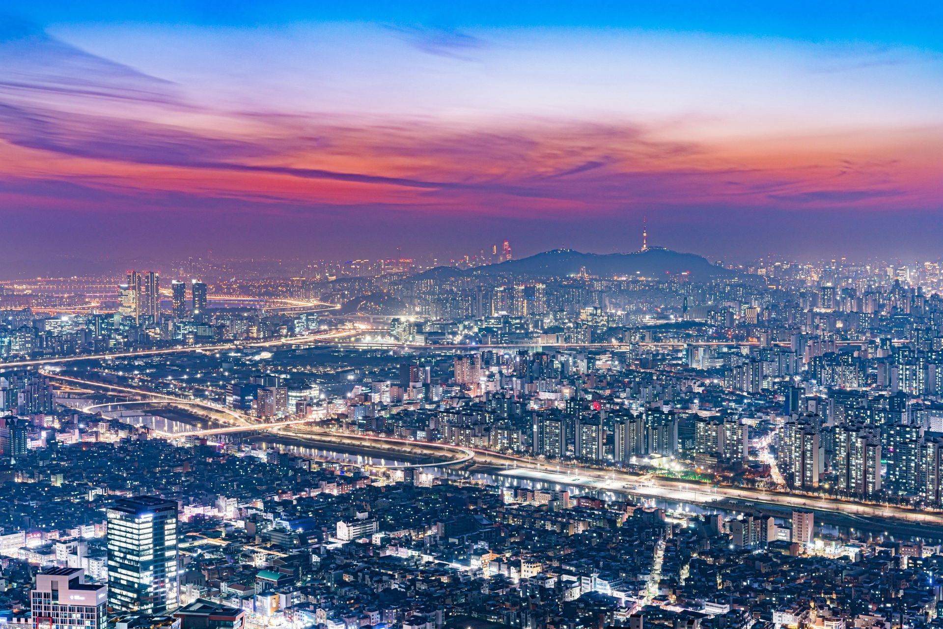 City skyline at dusk with glowing buildings and a pink-purple sunset over the hills