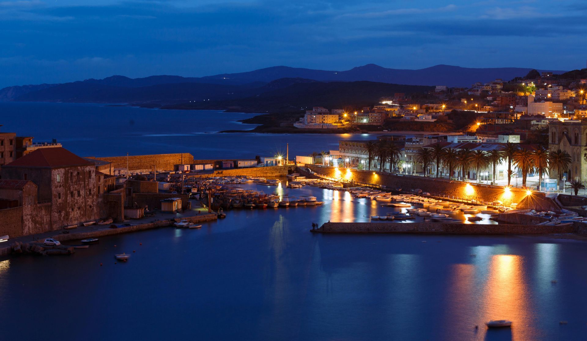 Blue-hour coastal harbor with lit buildings, calm water, and city lights reflecting at night