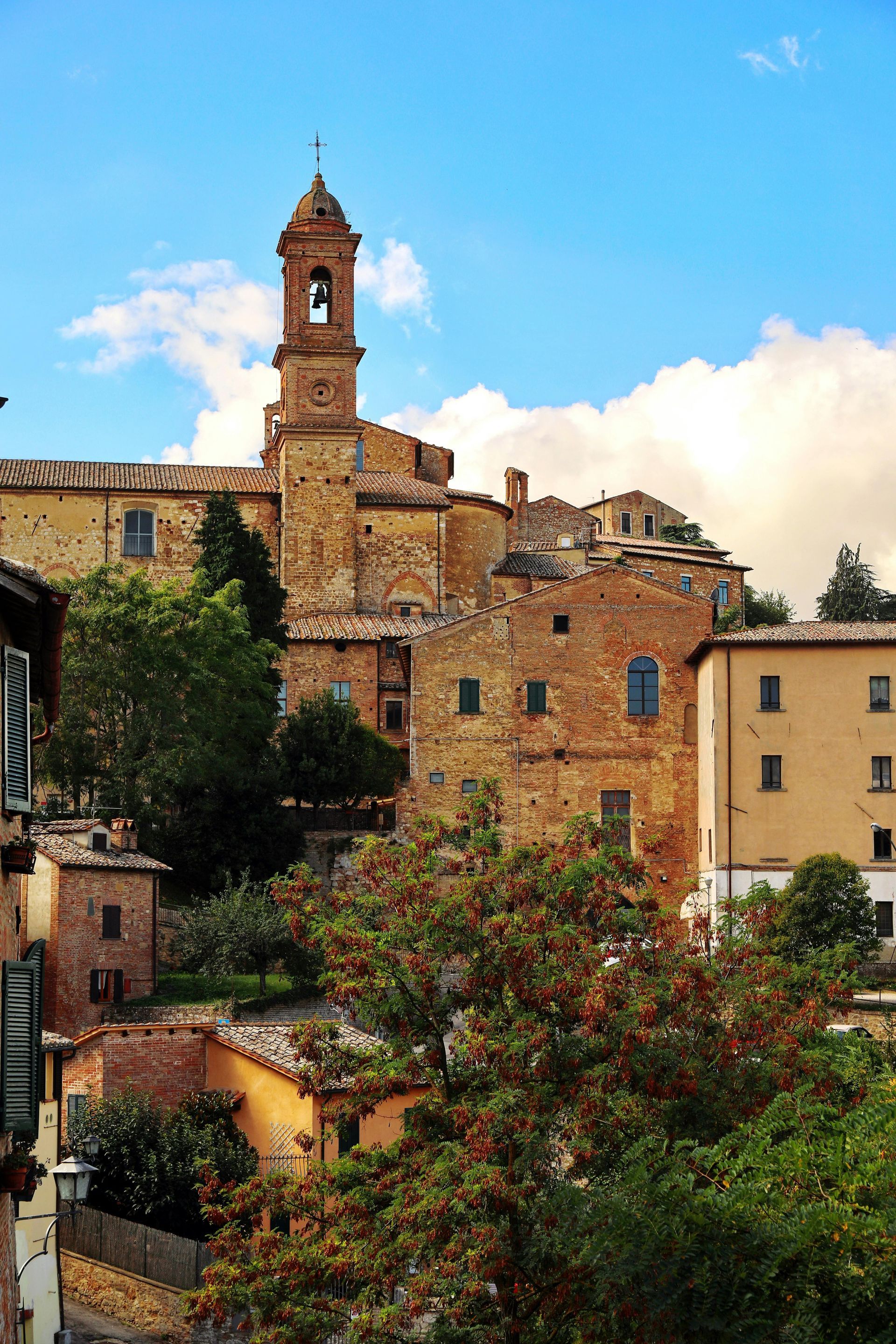 Brick buildings and a bell tower in an Italian town, beneath a blue sky with clouds.