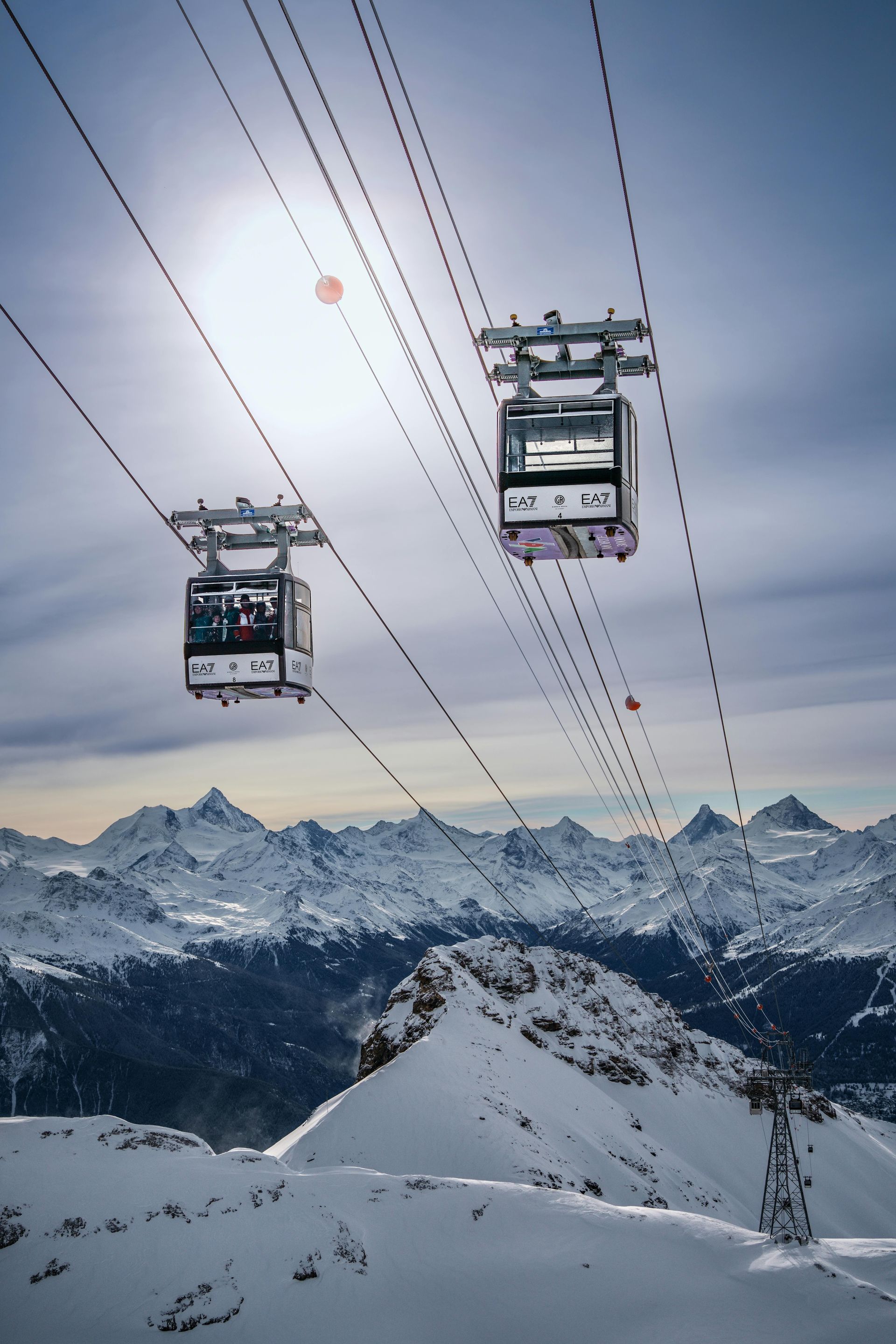 Two cable cars ascend over snow-covered mountains under a bright sun. Crans-Montana