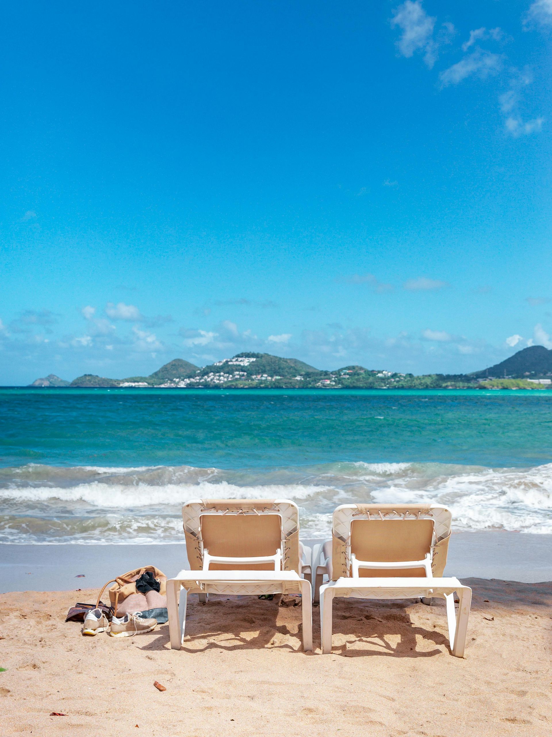 Two empty lounge chairs sit on a sandy beach facing the blue ocean and distant hills under a bright sky.