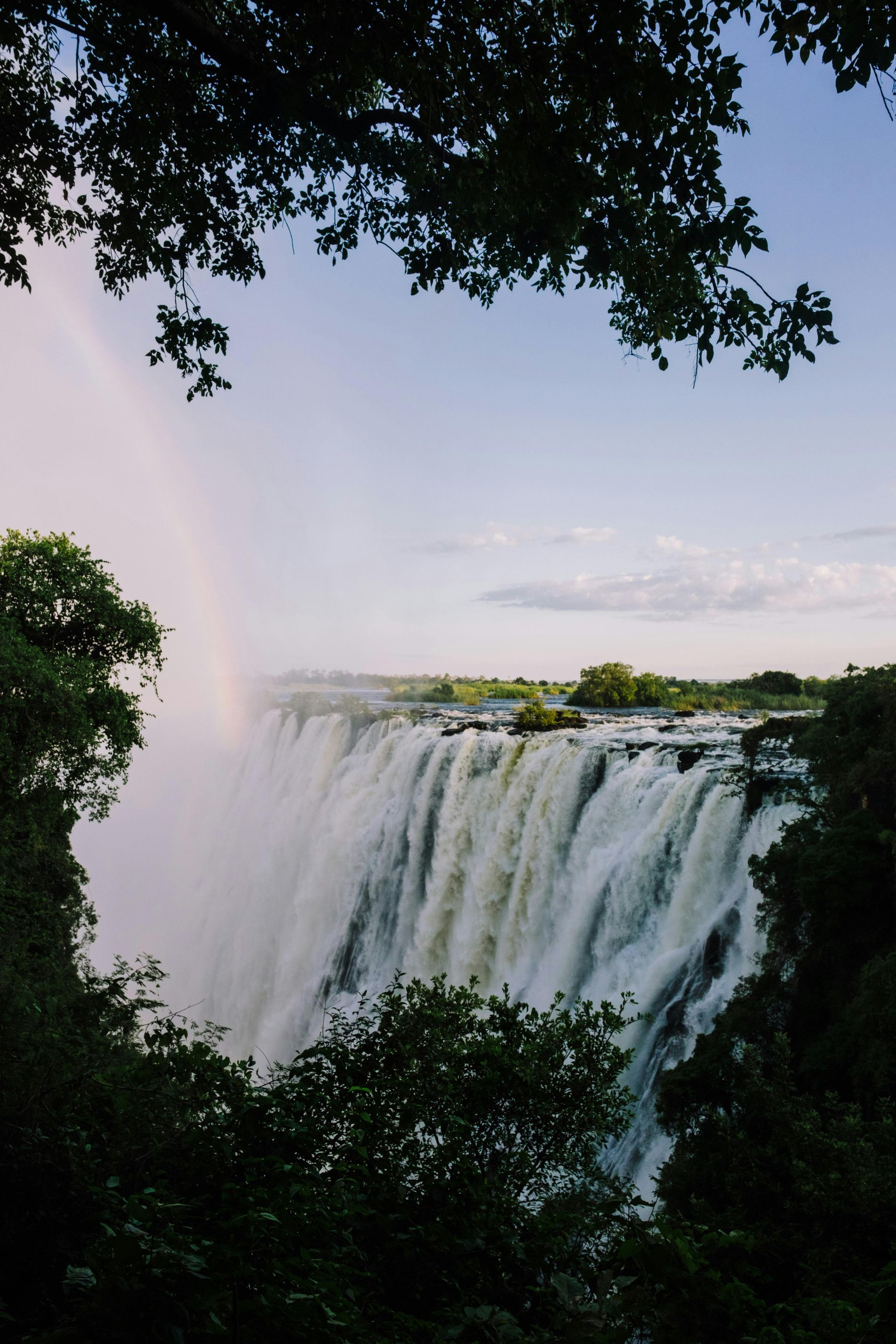 Victoria Falls cascading over a cliff, a rainbow arcs, seen through trees. Zimbabwe and Zambia – Victoria Falls and the Zambezi