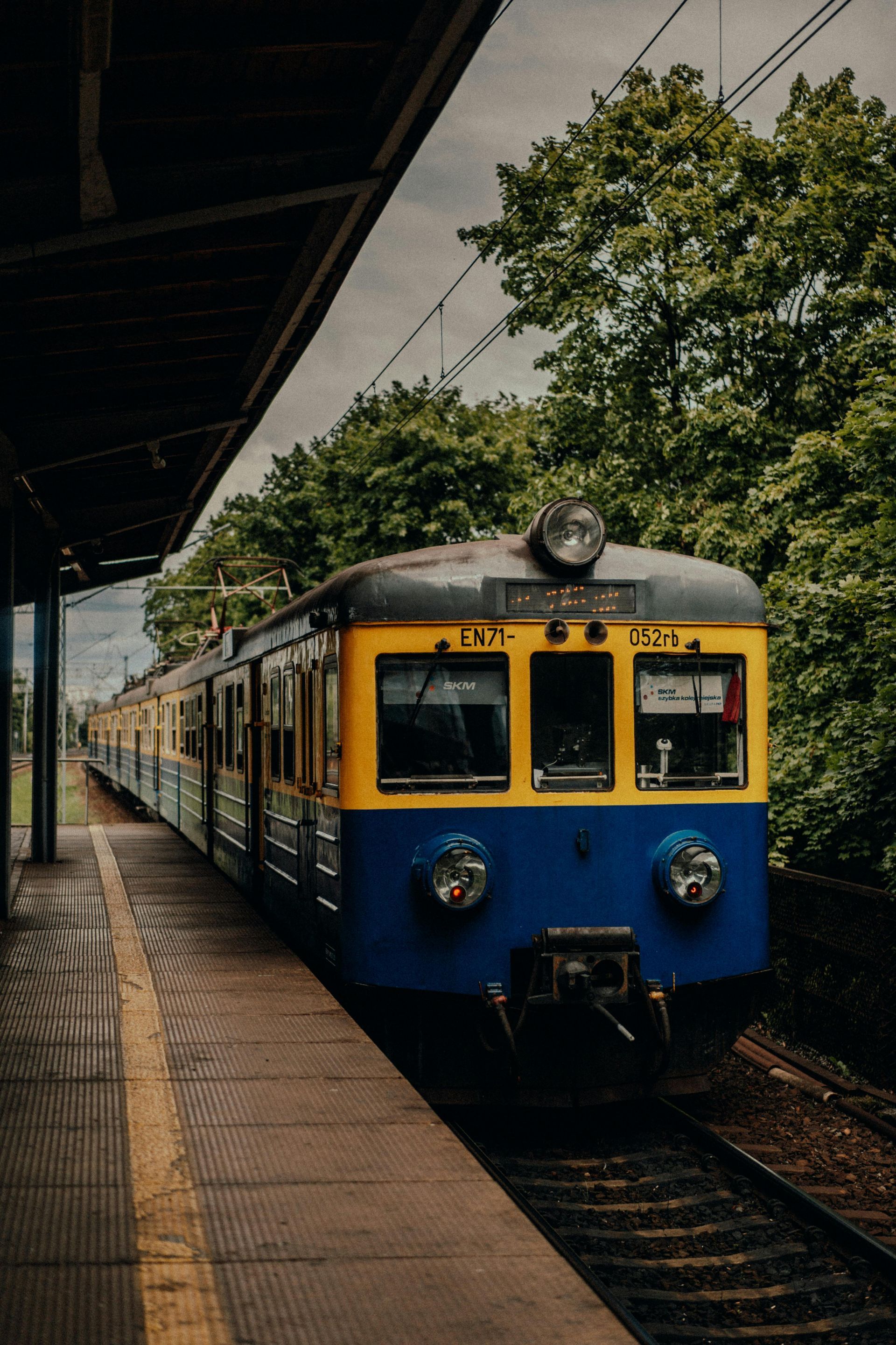 A blue and yellow passenger train stopped at a station platform next to green trees on an overcast day.