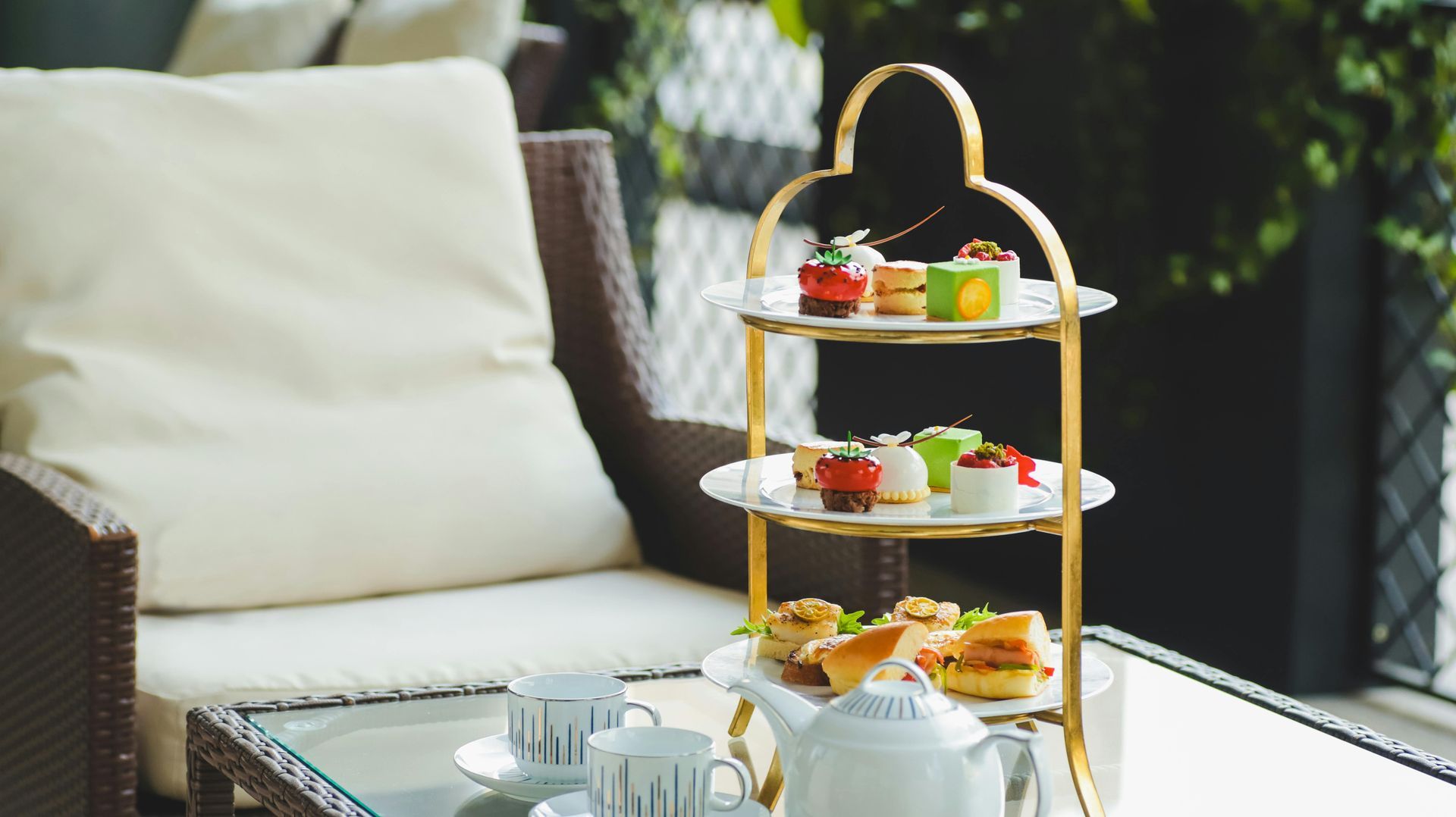 Afternoon tea set on a three-tiered stand; white teacups, teapot, and snacks beside a patio chair.
