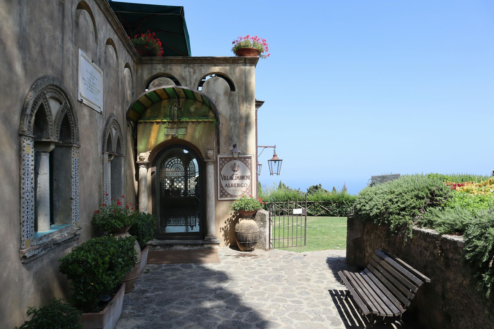 Old stone building with arched doorway, flowers, and a bench overlooking the sea. Ravello