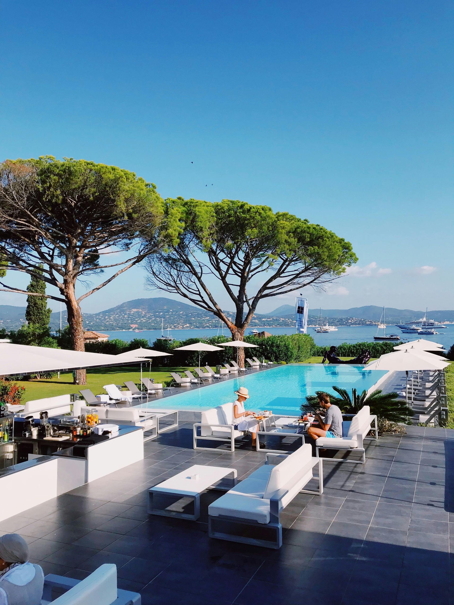 Poolside lounge area with a view of the ocean and mountains on a sunny day. French Riviera