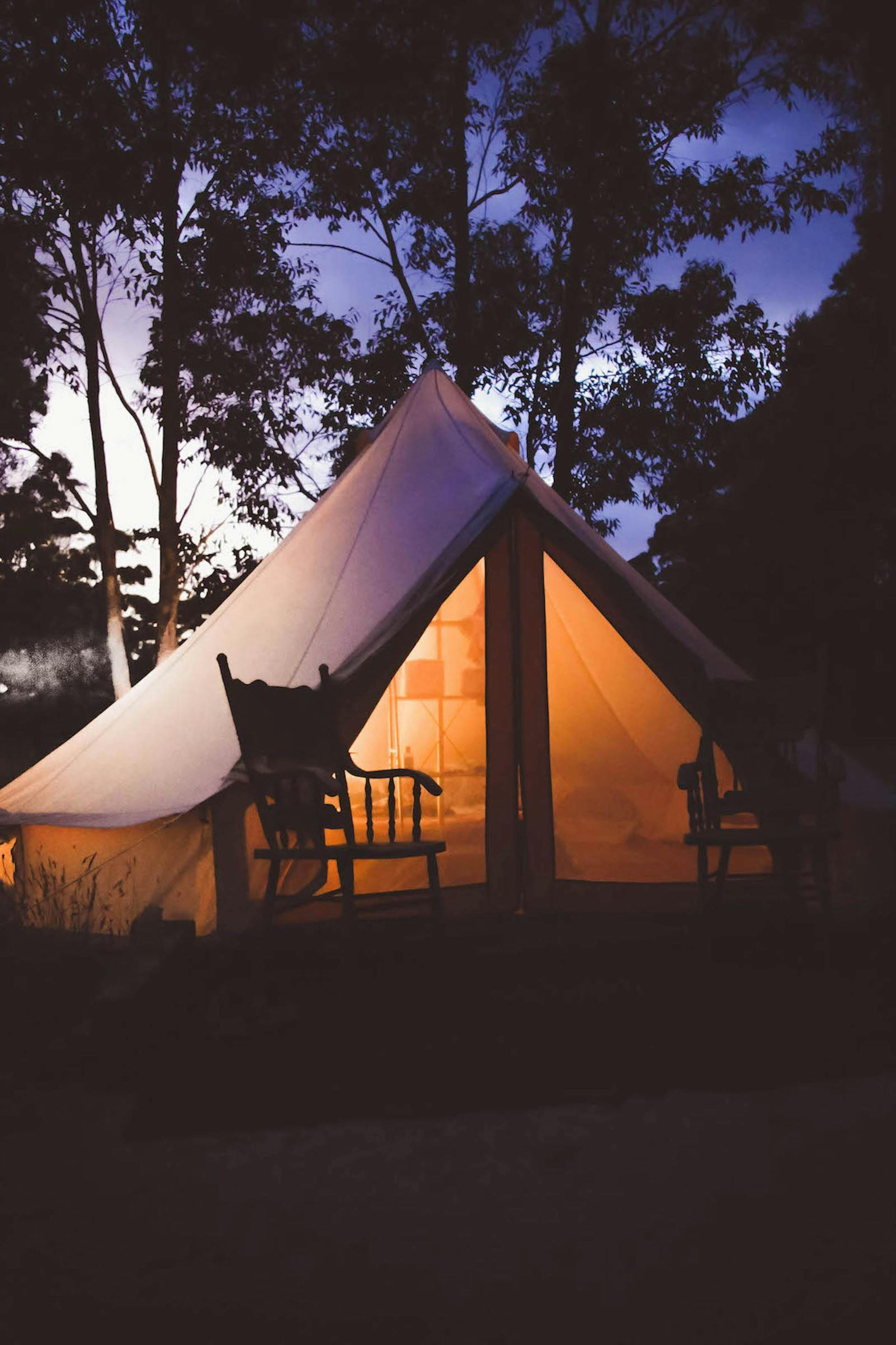 White tent lit from within, two chairs sit outside at dusk, trees in background.