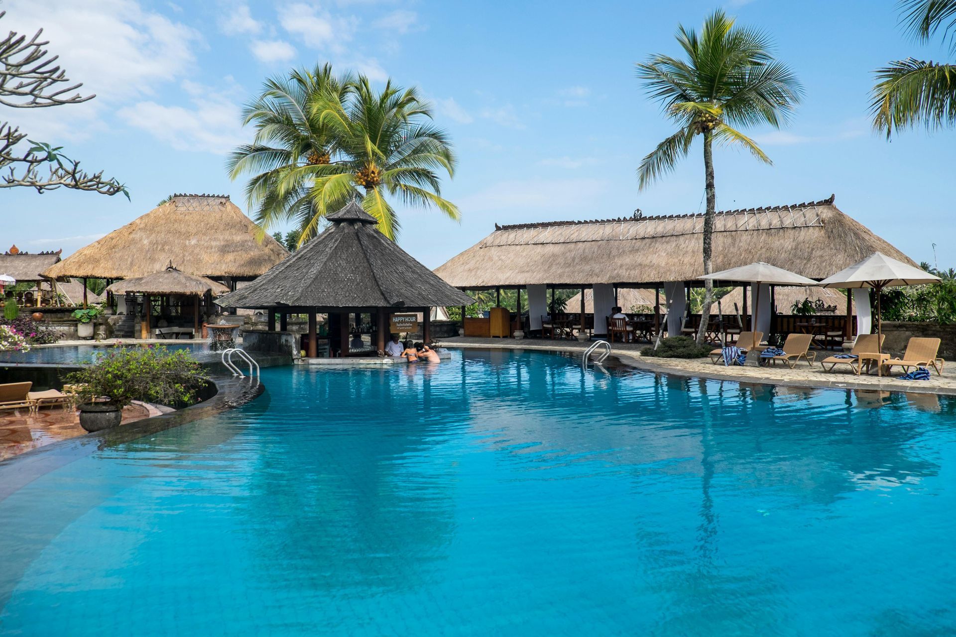 Swimming pool at a resort with thatched roof structures, palm trees, and blue sky.