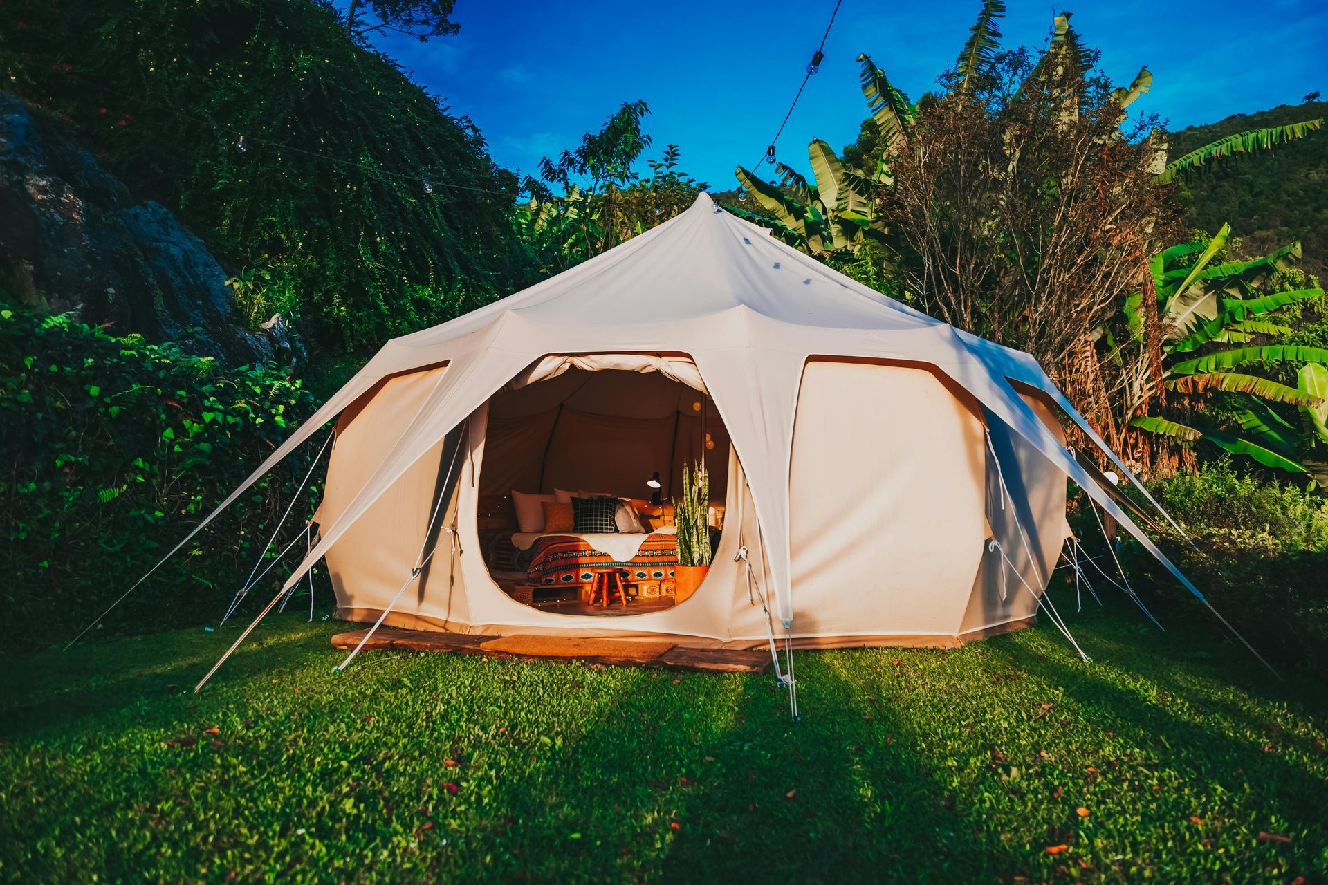 Beige glamping tent on green grass, open to interior with furniture, surrounded by lush greenery.