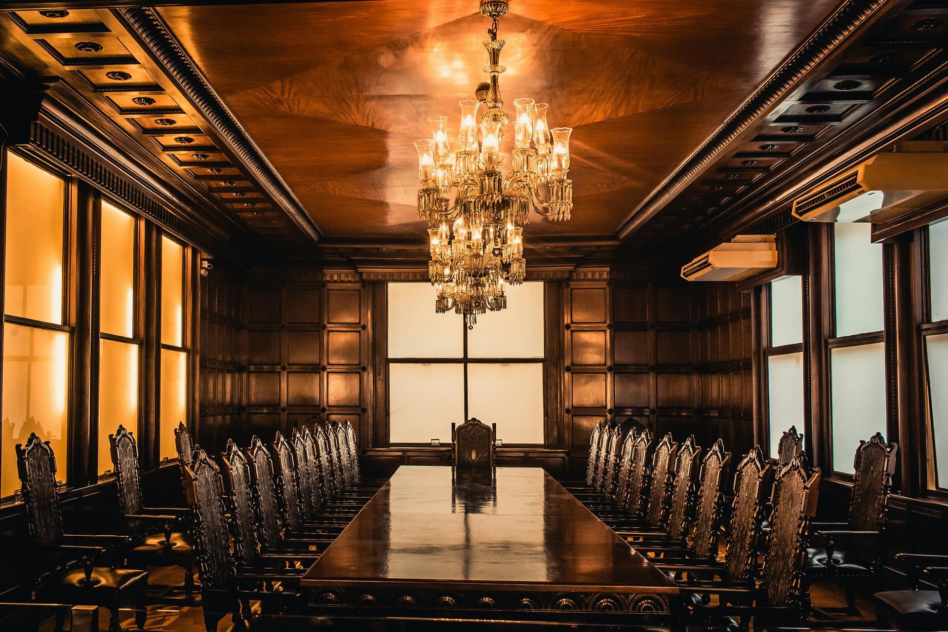 A grand, wooden dining room with long table, ornate chairs, chandelier, and large windows.