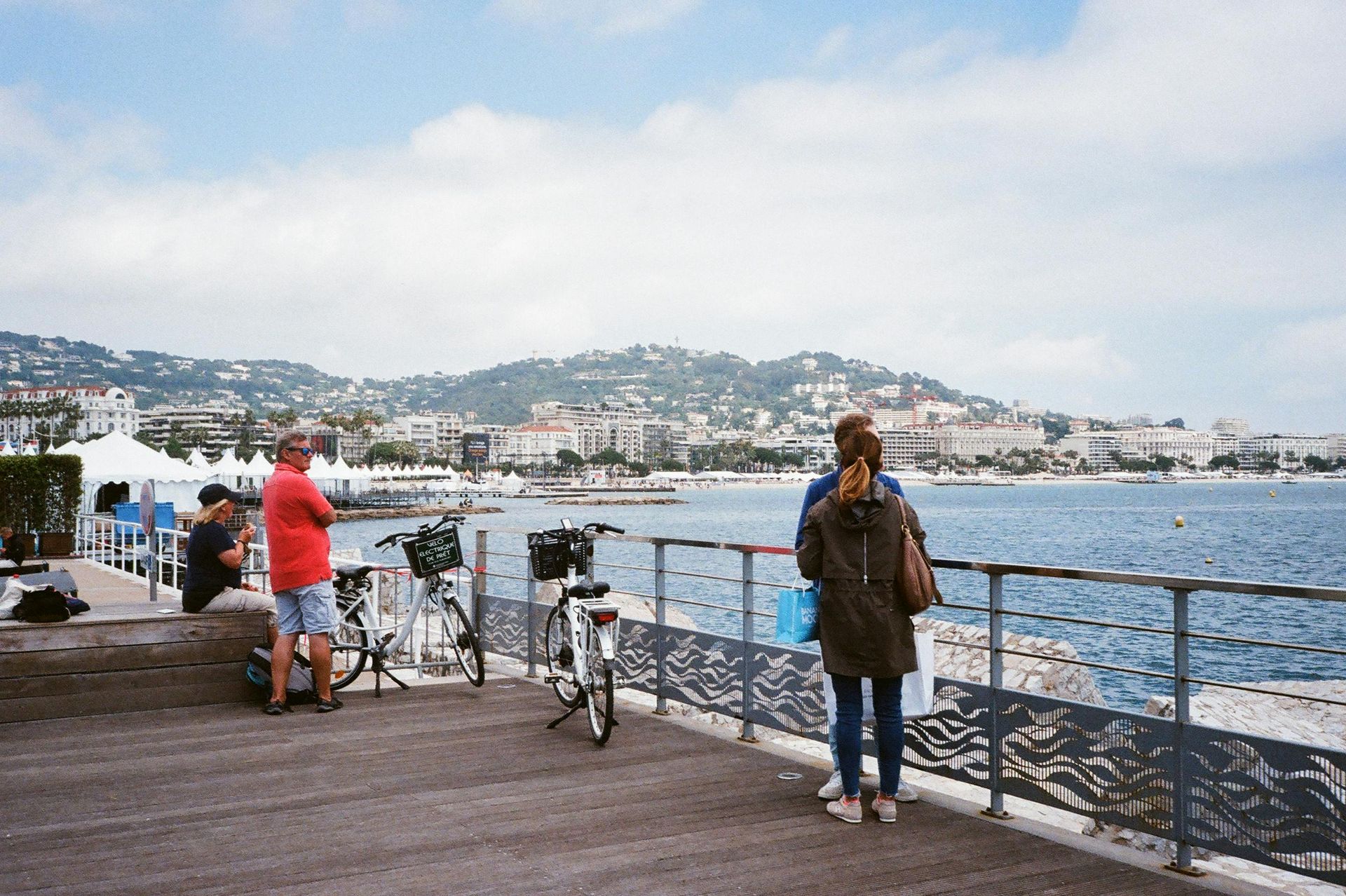People on a seaside promenade looking at a city on the coast. Blue water, clear sky. Cannes & Cap d’Antibes