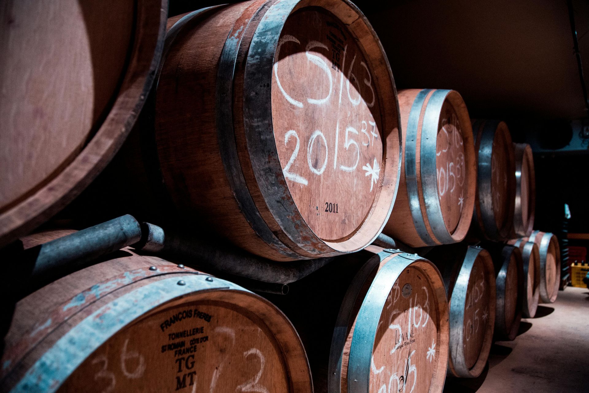 Wooden wine barrels stacked in a dim cellar, featuring handwritten chalk markings and metal rings.