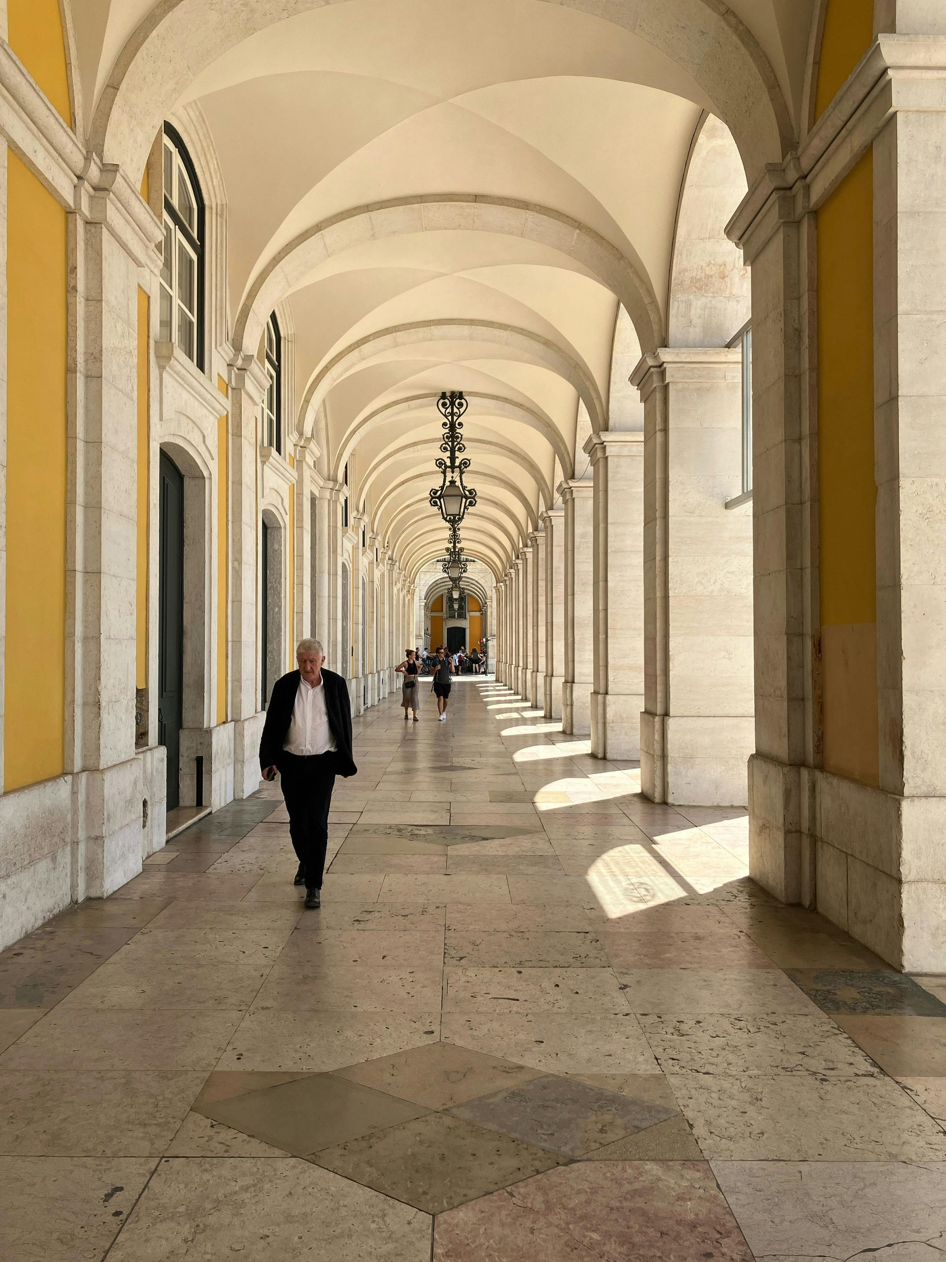 Arcaded corridor with repeating arches and a few people walking in sunlight.
