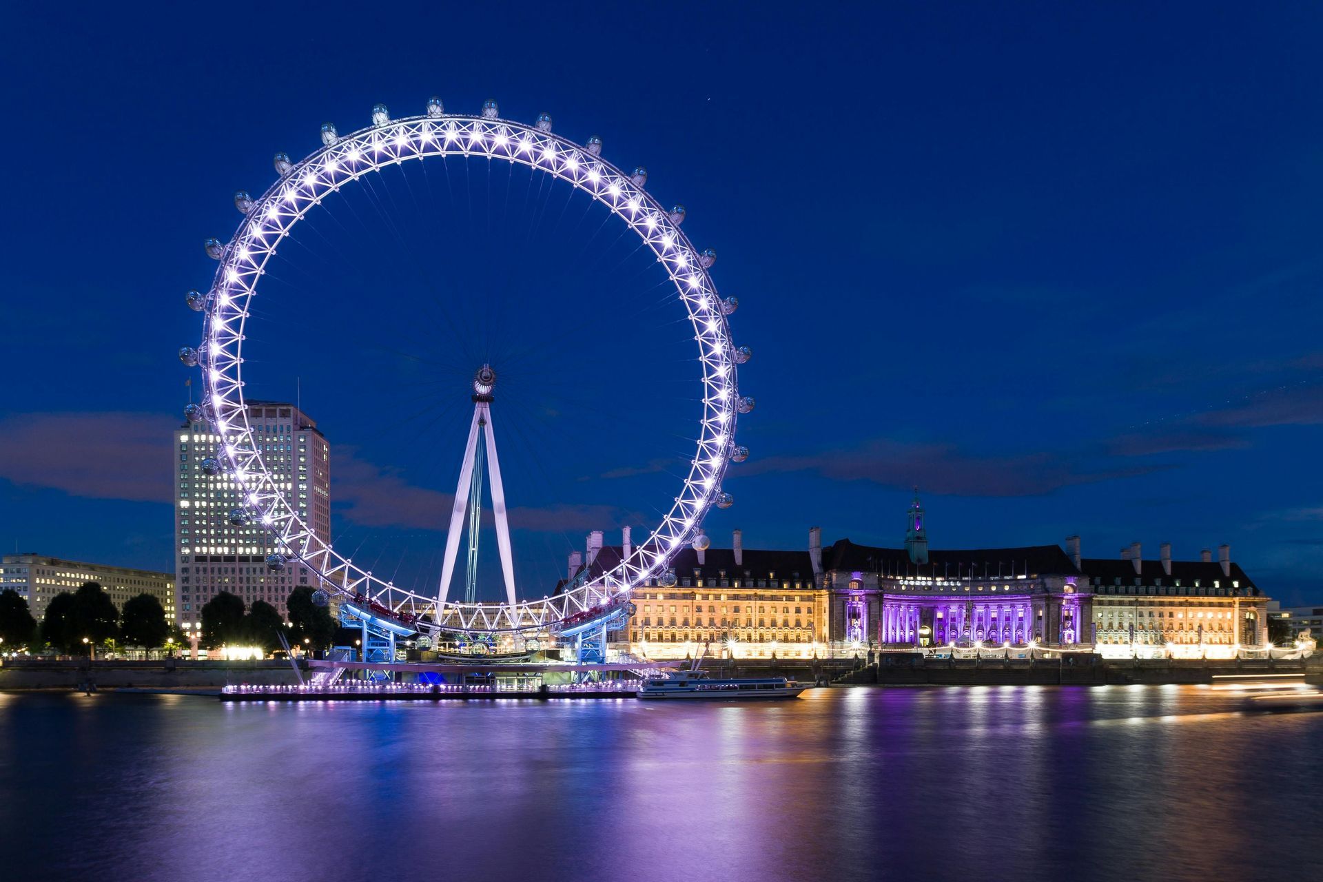 London Eye illuminated at night, reflecting in the Thames. Buildings line the river, blue sky.
