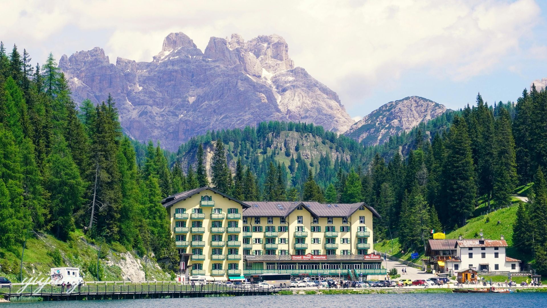 Hotel by a lake with mountains in the background. Green building with pine trees surrounding.