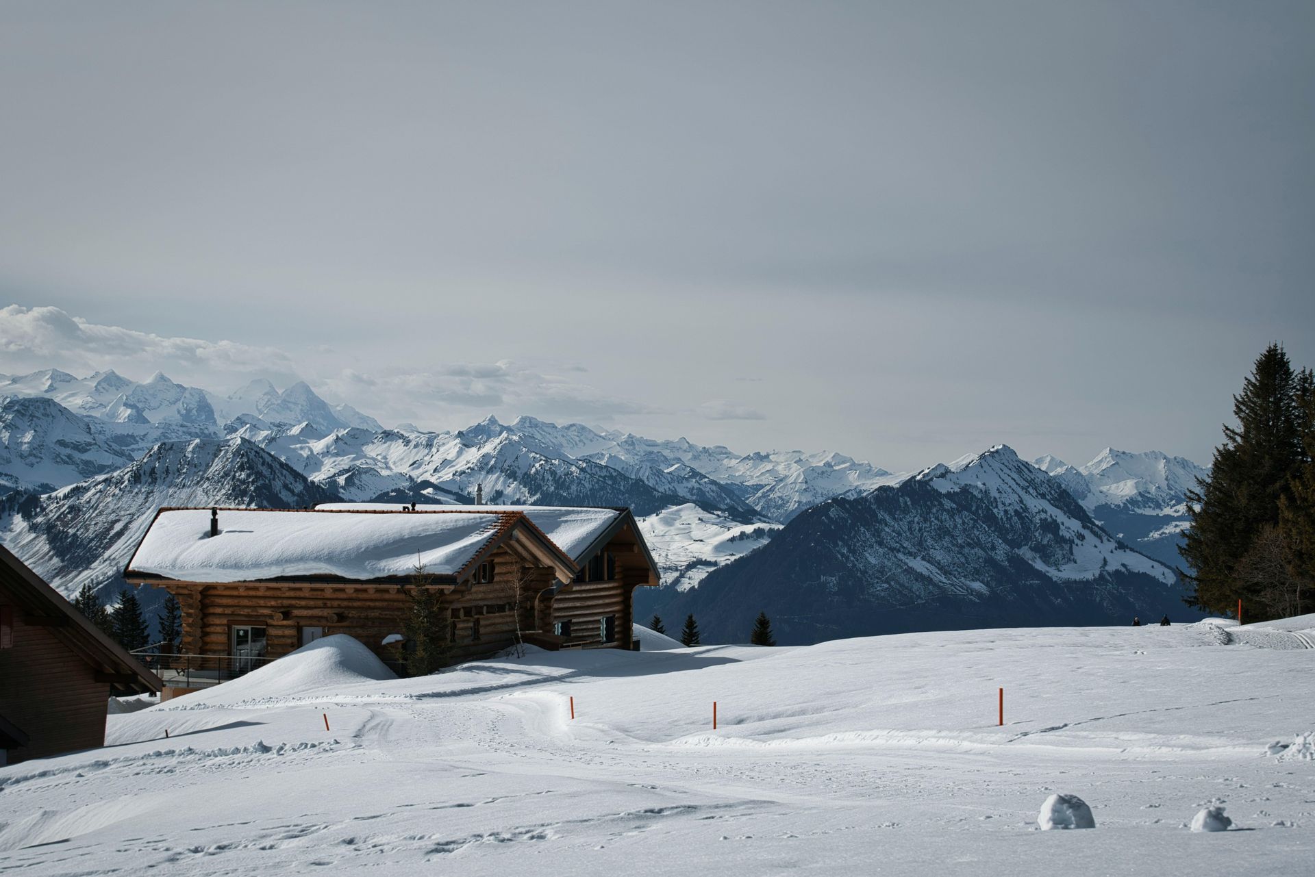 Snow-covered wooden cabins on a snowy mountain with a backdrop of snow-capped mountain ranges under a gray sky. Gstaad