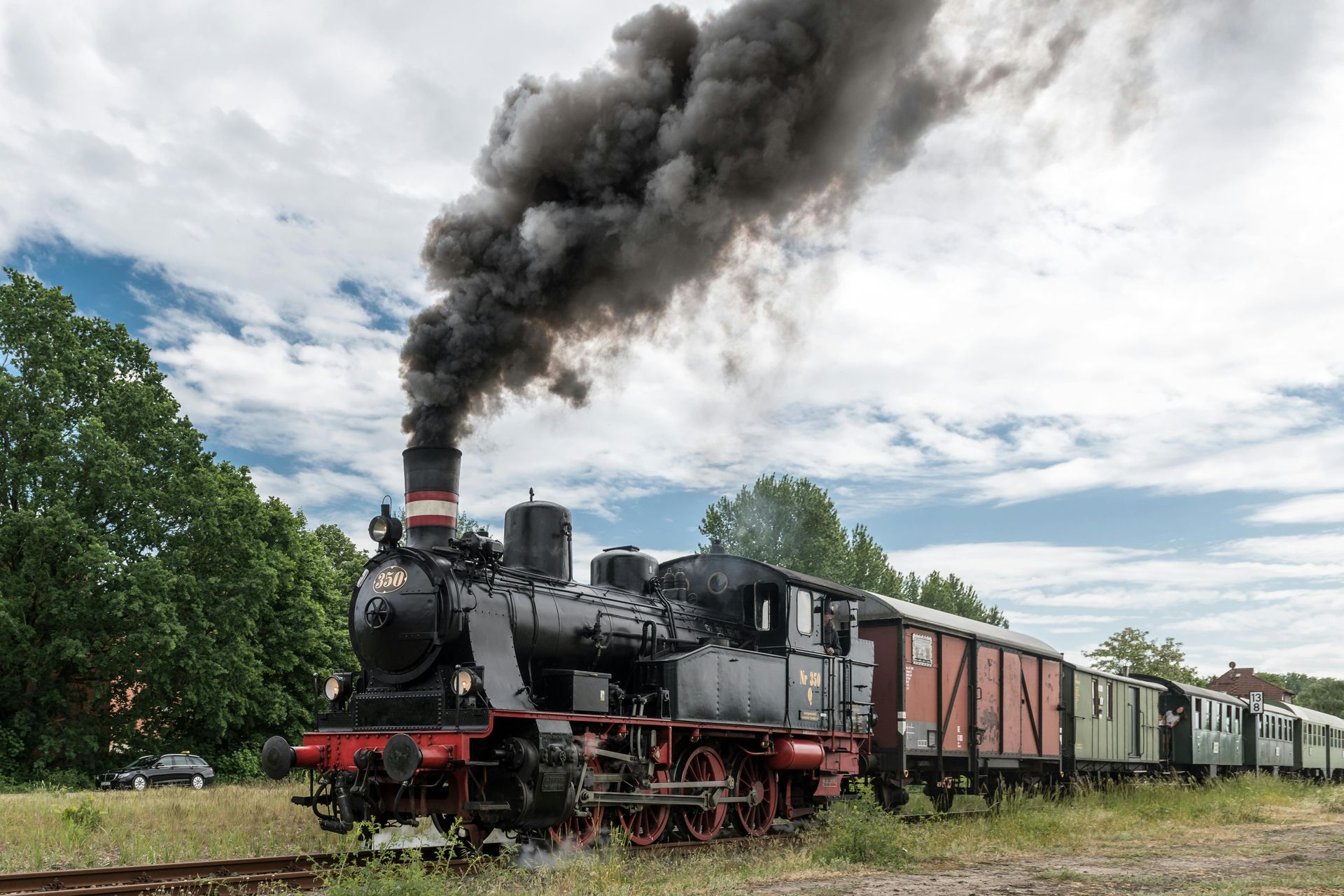 A black steam locomotive pulls vintage train cars through a grassy field under a cloudy sky, emitting thick dark smoke.