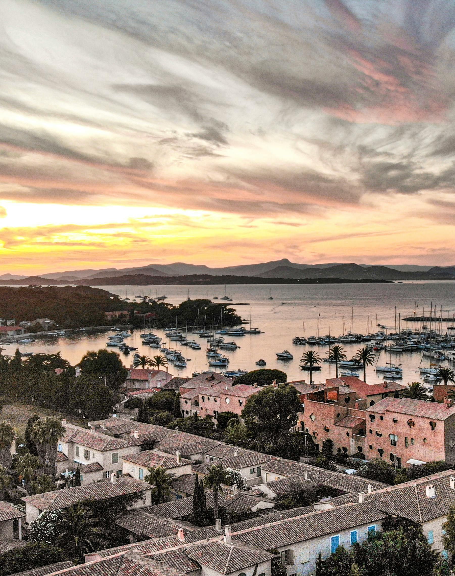 Sunset over harbor with boats, buildings, and mountains. Orange and pink sky. Saint-Tropez