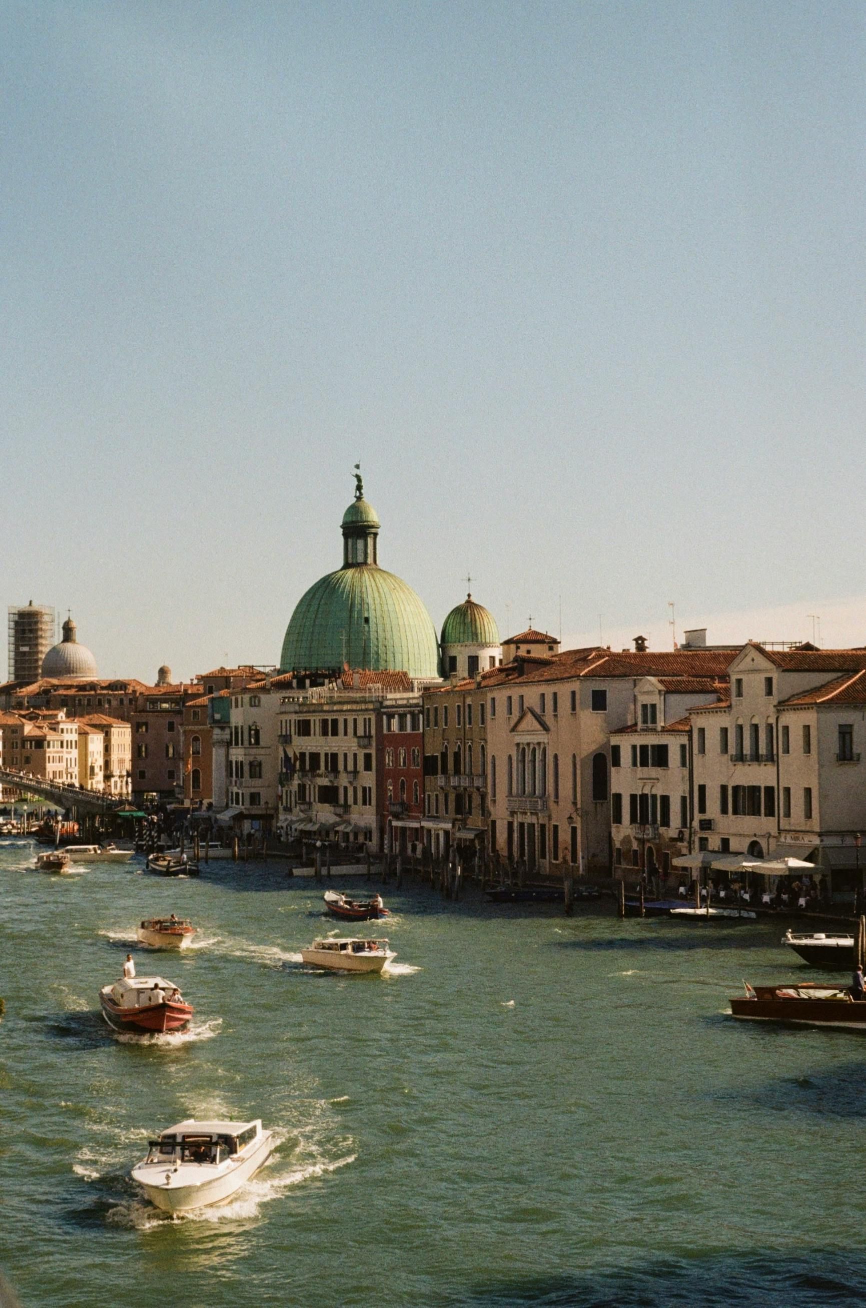 Venice canal scene with boats, buildings, and a green dome under a clear blue sky.