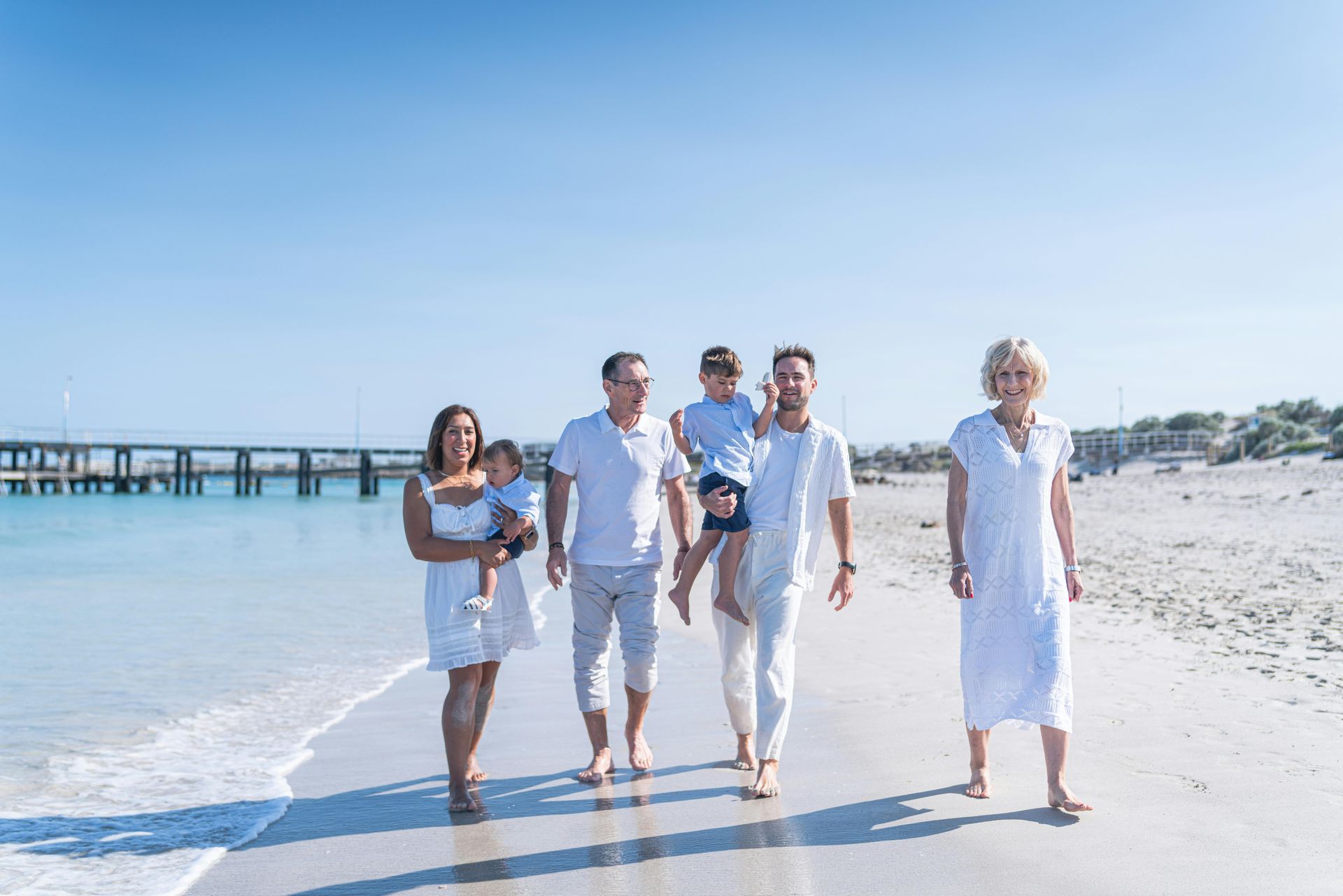 A family dressed in white walks along a sandy beach near a pier under a bright, clear blue sky.