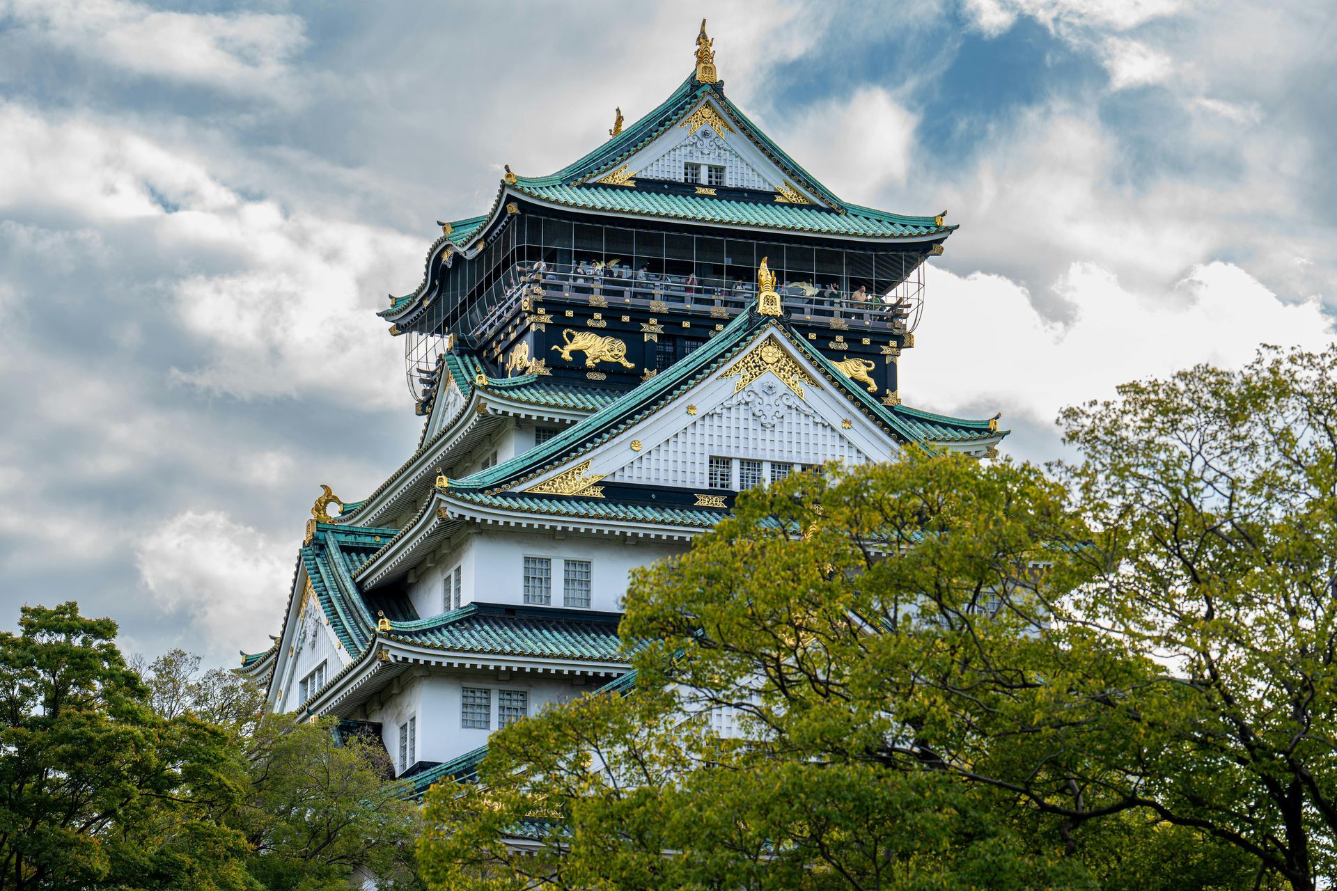 Osaka Castle with white walls, green roofs, and gold accents, nestled behind vibrant green trees under a cloudy sky.