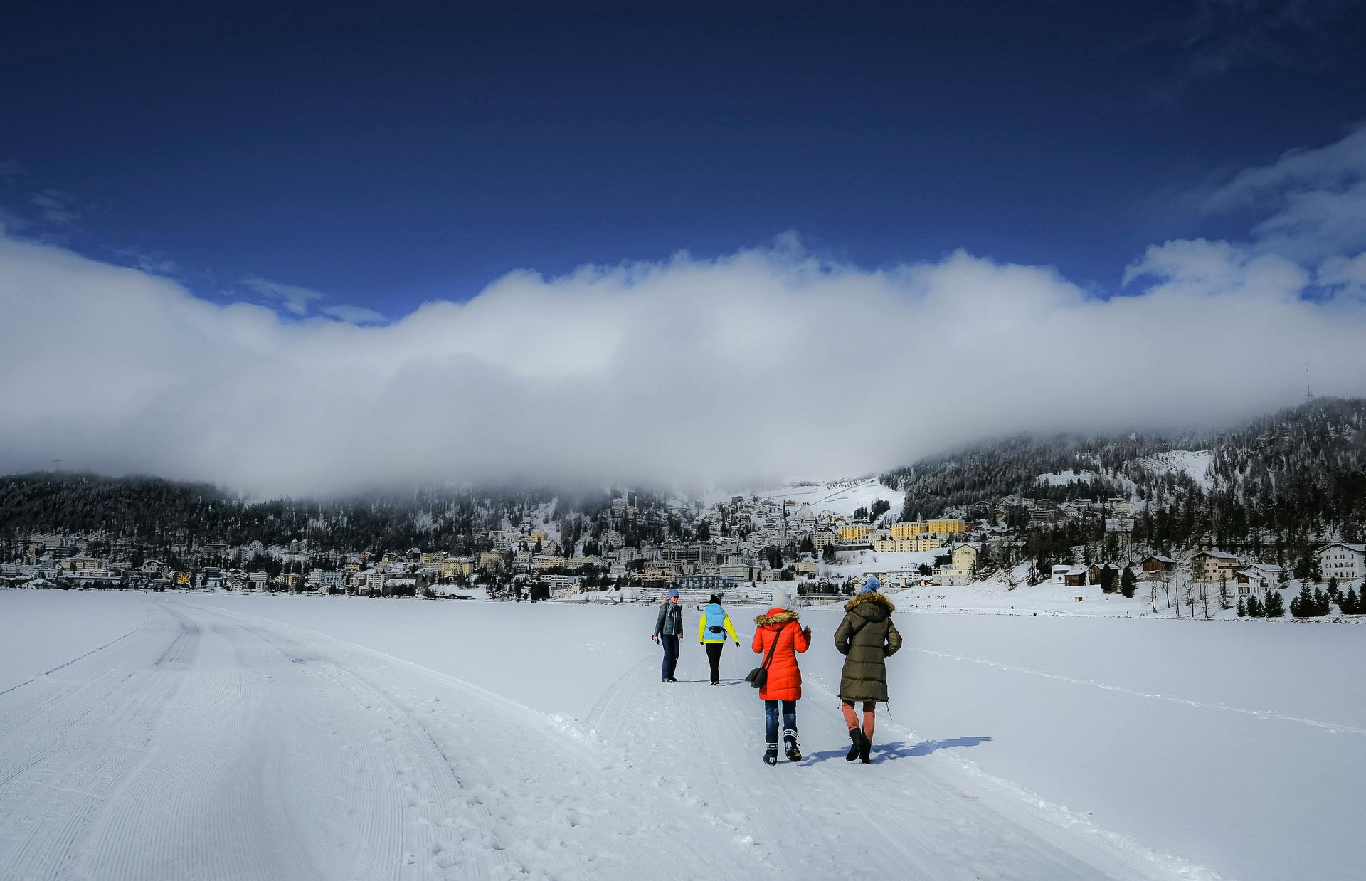 People walking on snow-covered ground towards a village beneath a cloudy sky. Brightly colored coats stand out. St. Moritz