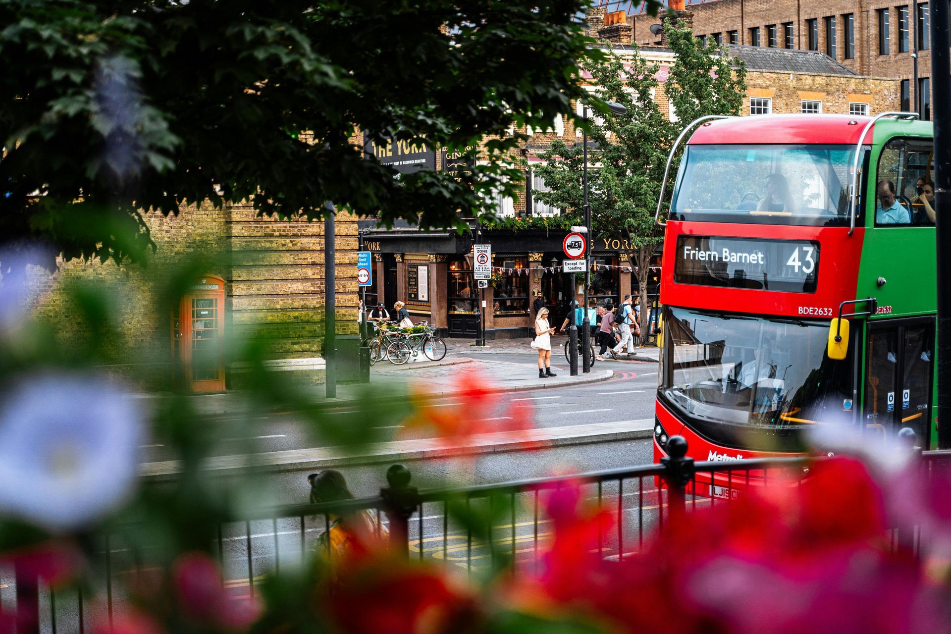 Red and green double-decker bus on a street; people and shops in the background; colorful flowers in the foreground.