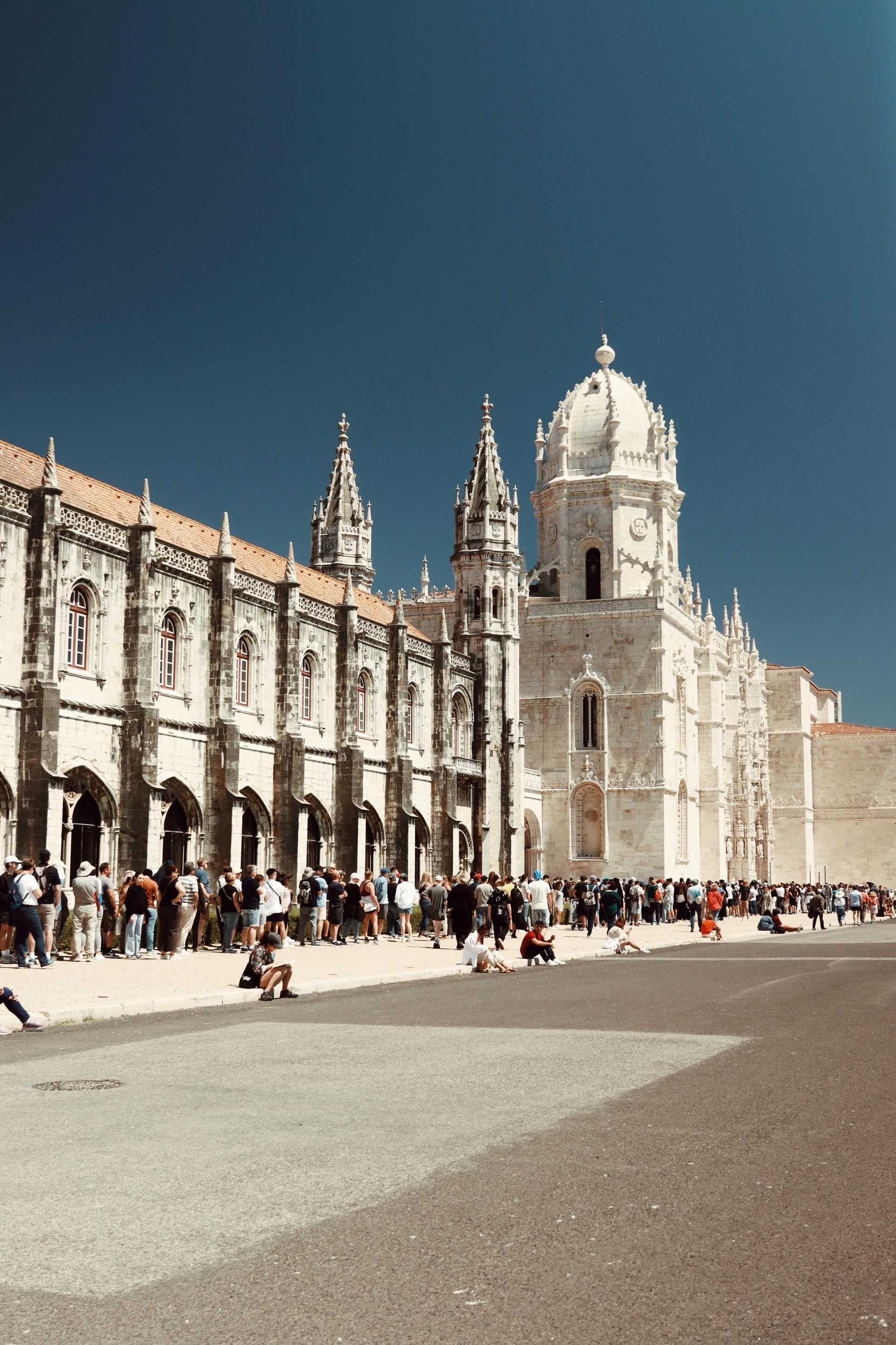 Crowd in a historic plaza beside ornate white cathedral and bell towers under a blue sky. Lisbon