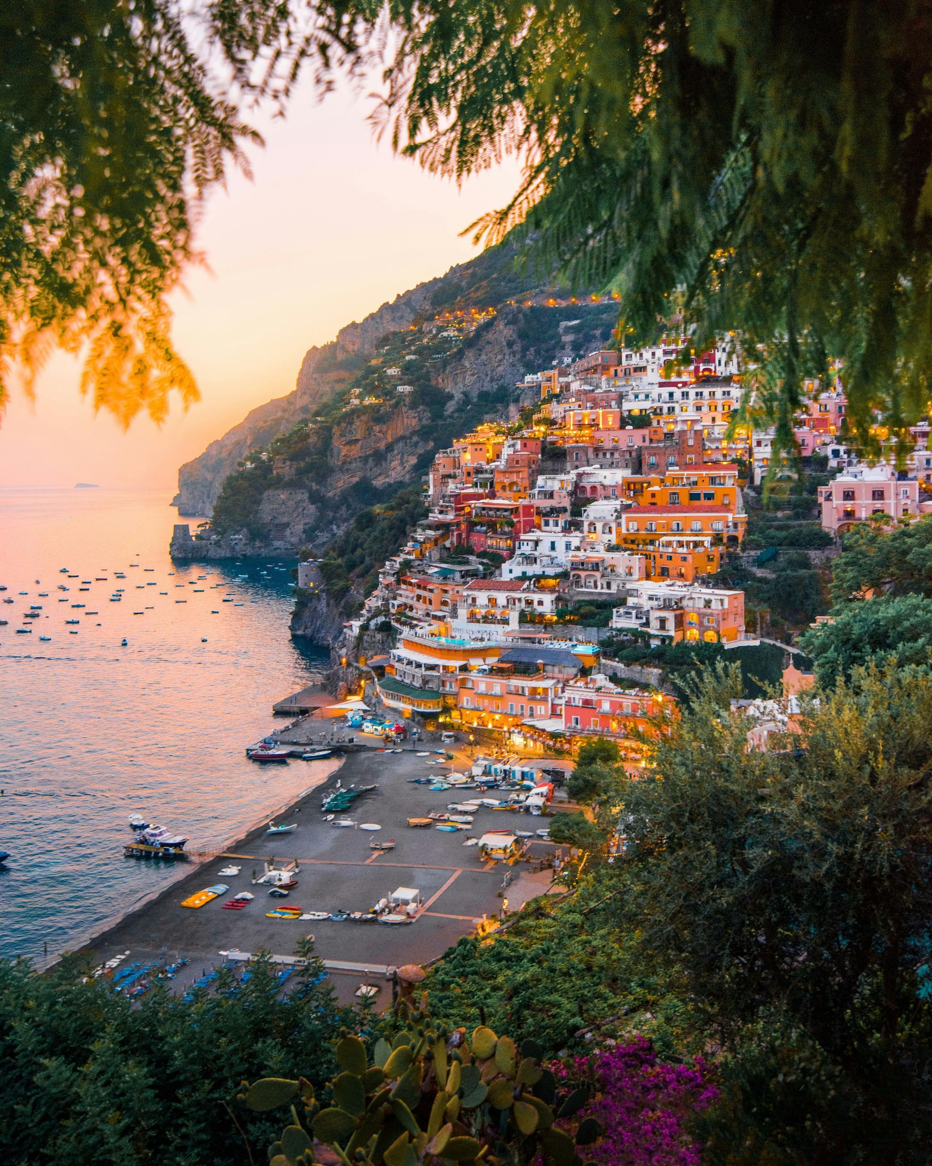 Coastal Italian town at sunset: colorful buildings on a cliffside, boats in the sea, framed by foliage. Positano Amalfi