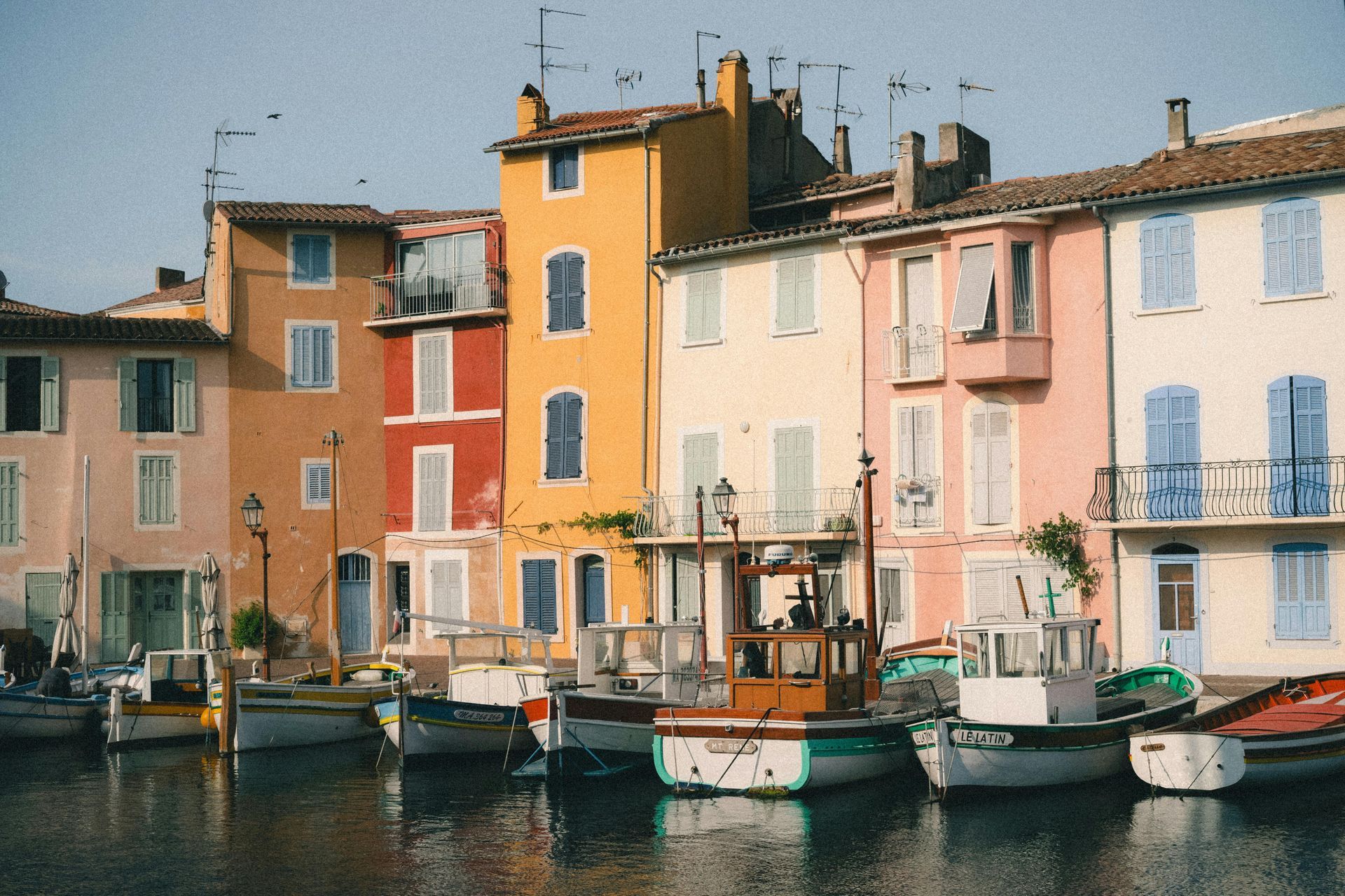 Colorful buildings and boats line a calm harbor; pastel facades, blue shutters, and reflections in the water.